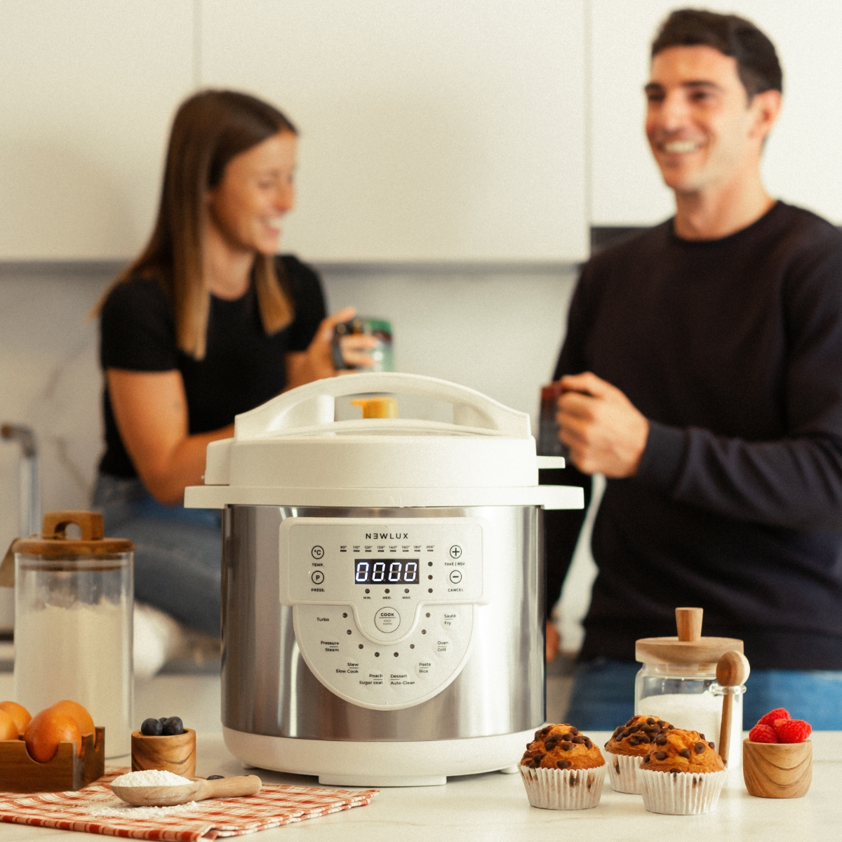 Una olla de cocción múltiple en una encimera con magdalenas, ingredientes y dos personas al fondo.
