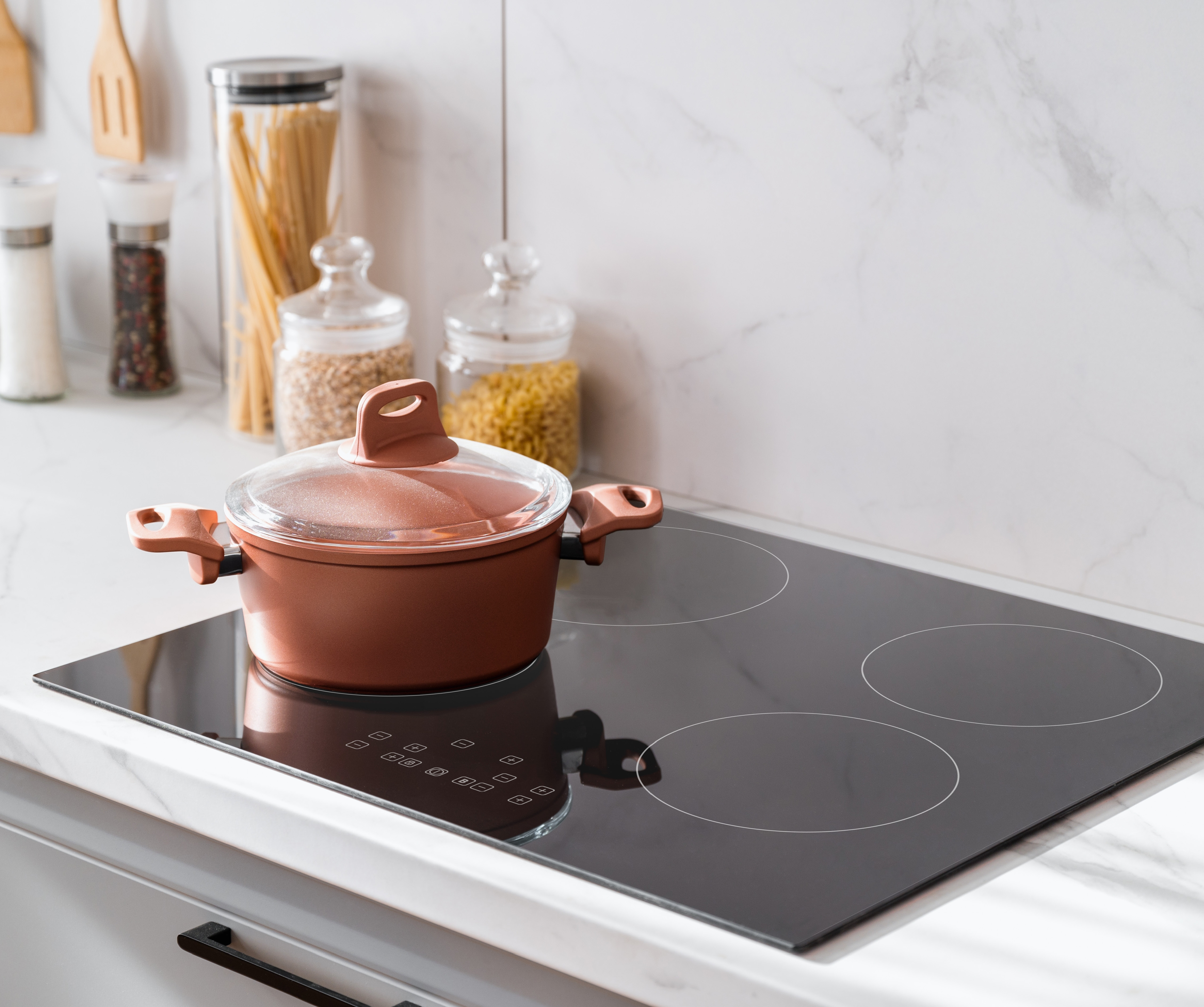 Kitchen scene with a pot on a black stovetop. White marble background. Wooden utensils in back.