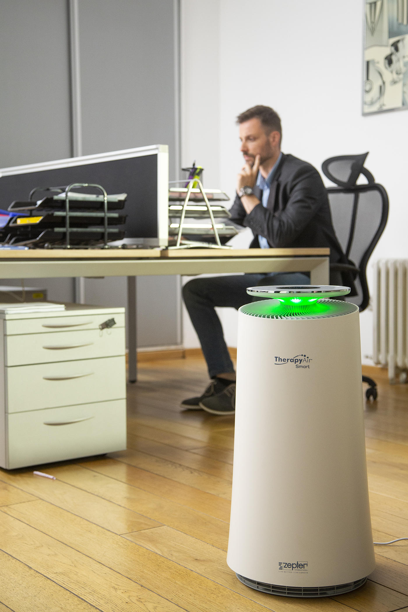 Ein Mann sitzt am Schreibtisch mit Luftreiniger und Laptop. Der Reiniger ist weiß mit grünem Licht, Laptop oben drauf.