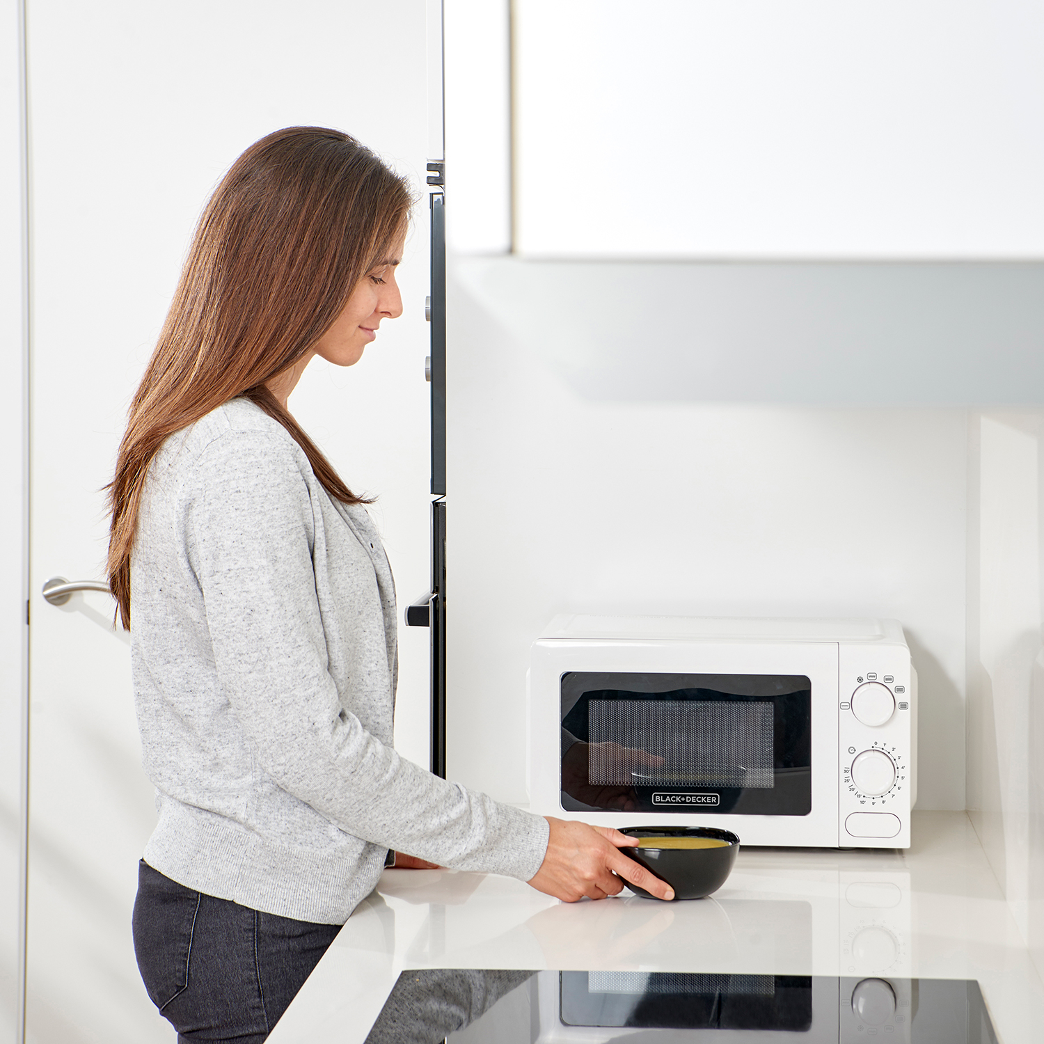 Una mujer con un suéter gris en una cocina, sacando un tazón del microondas.
