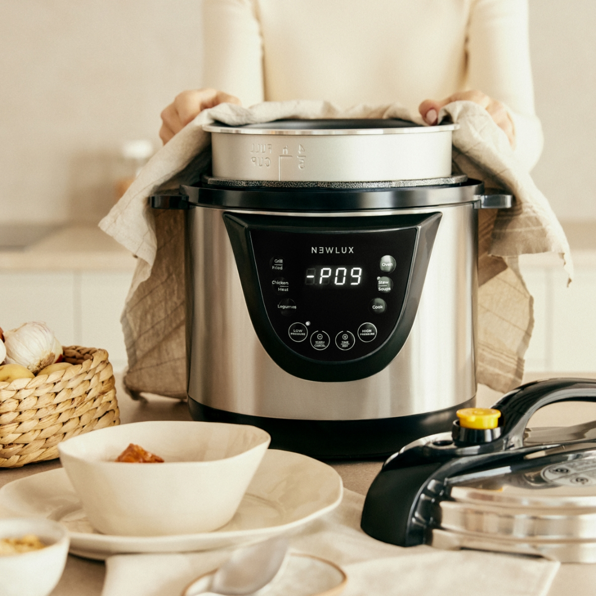 Una mujer sosteniendo una tapa sobre una olla a presión plateada, con otros elementos de cocina visibles.