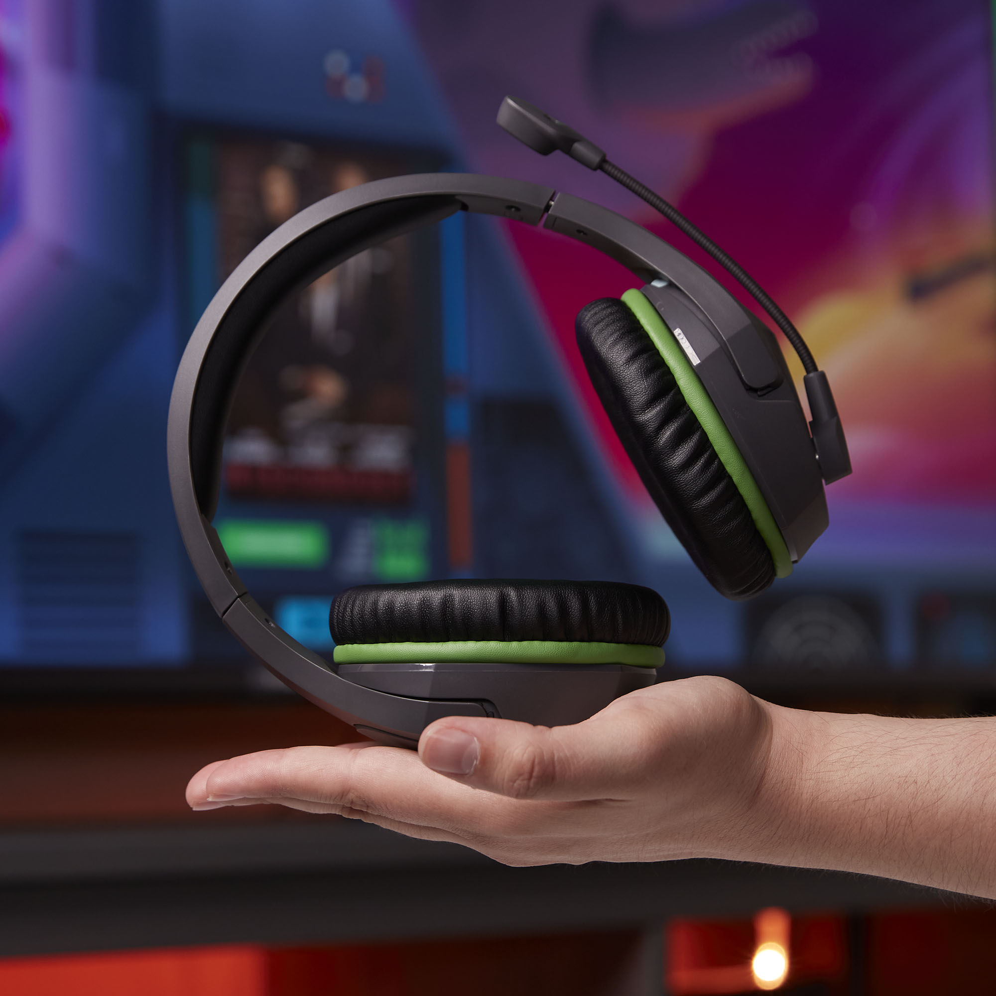 Hand hält ein Headset. Dunkelgraues und grünes Headset mit Mikrofon. Der Hintergrund ist ein verschwommener TV-Bildschirm.