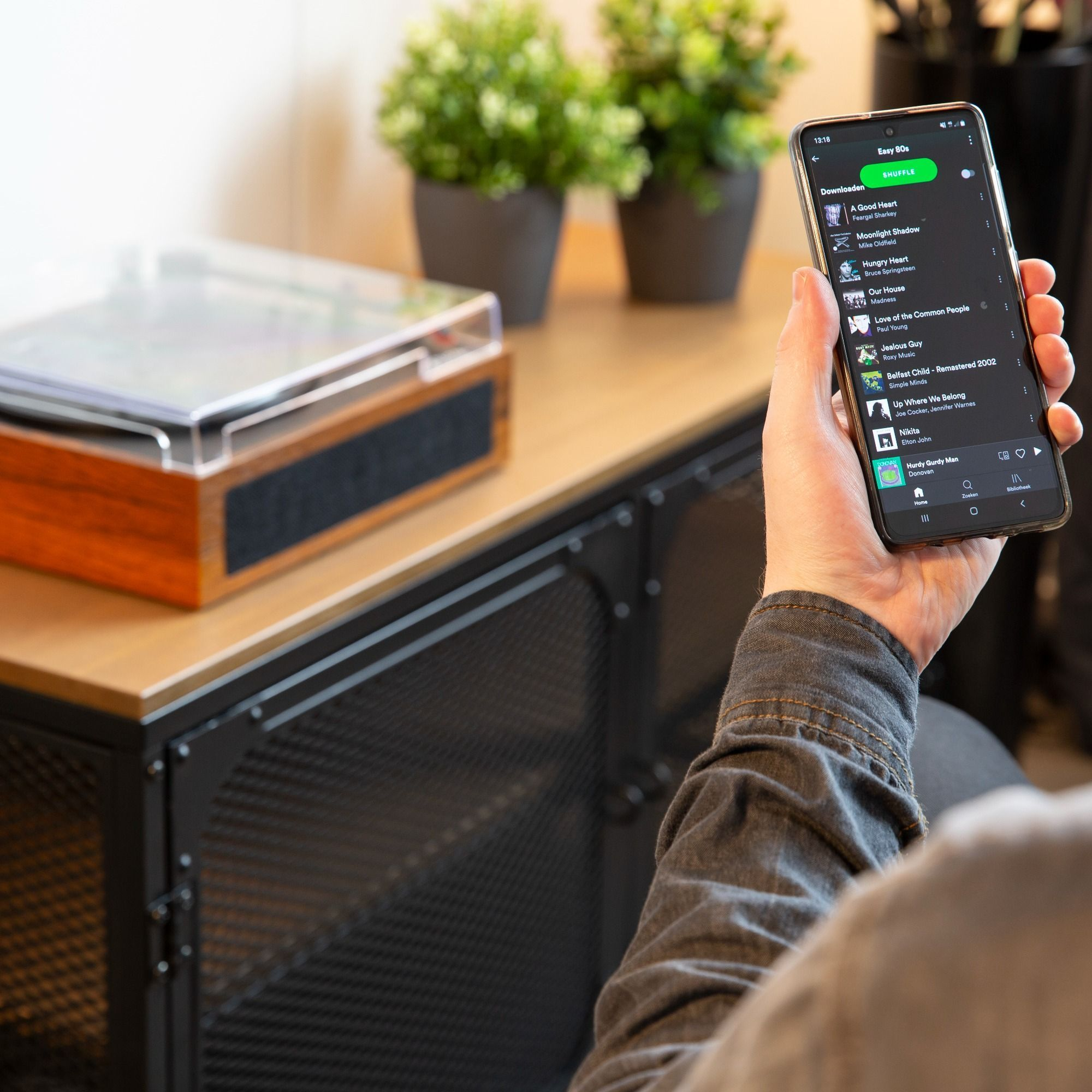 A person holds a phone with a music app. A record player and plants sit on a cabinet.