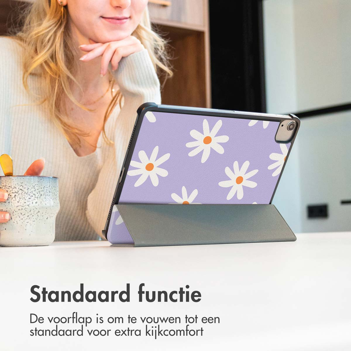 Een vrouw met een mok gebruikt een tablet met een bloemenhoes, op een witte tafel.