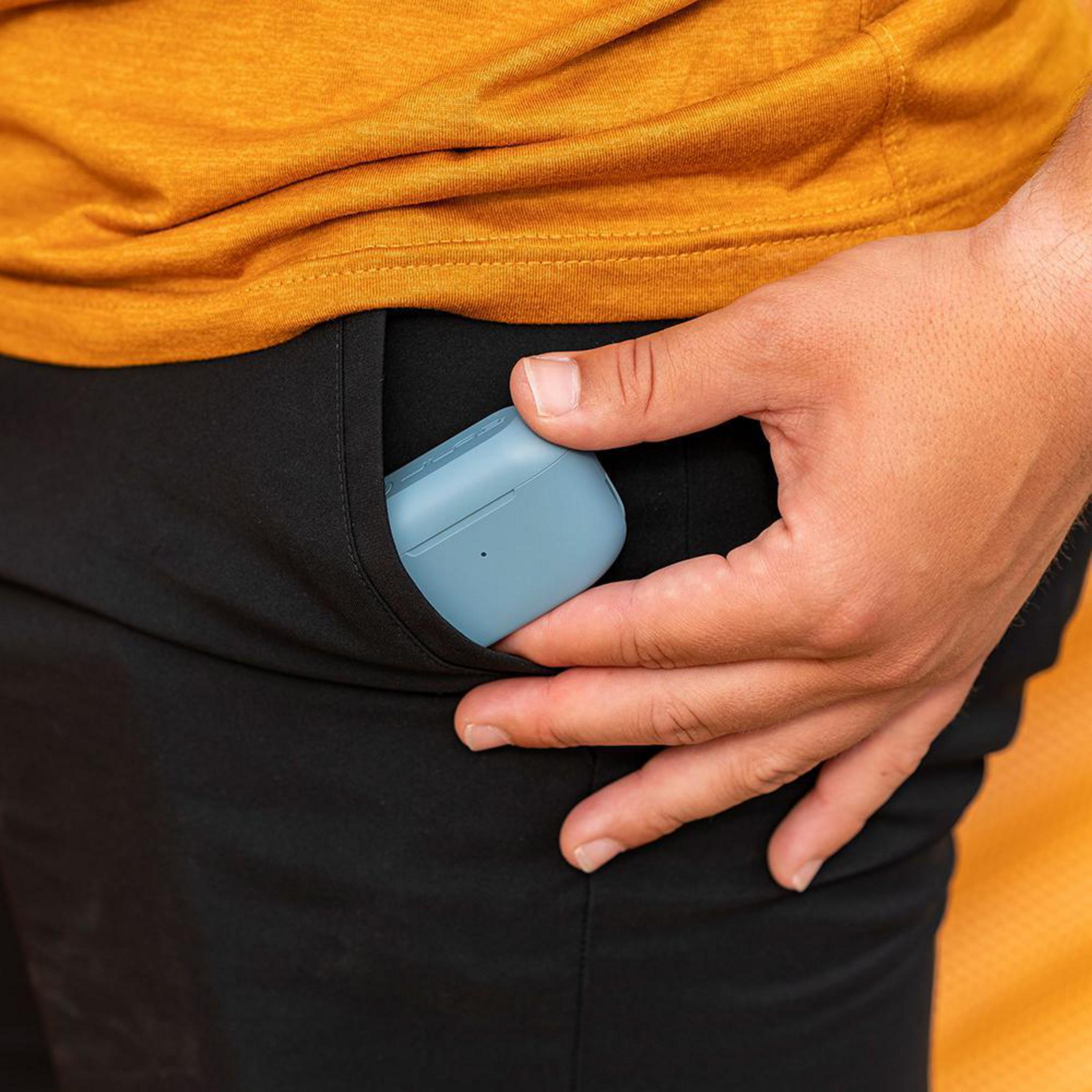 Dłoń w kieszeni trzymająca niebieskie etui na słuchawki. Osoba ma na sobie czarne szorty.