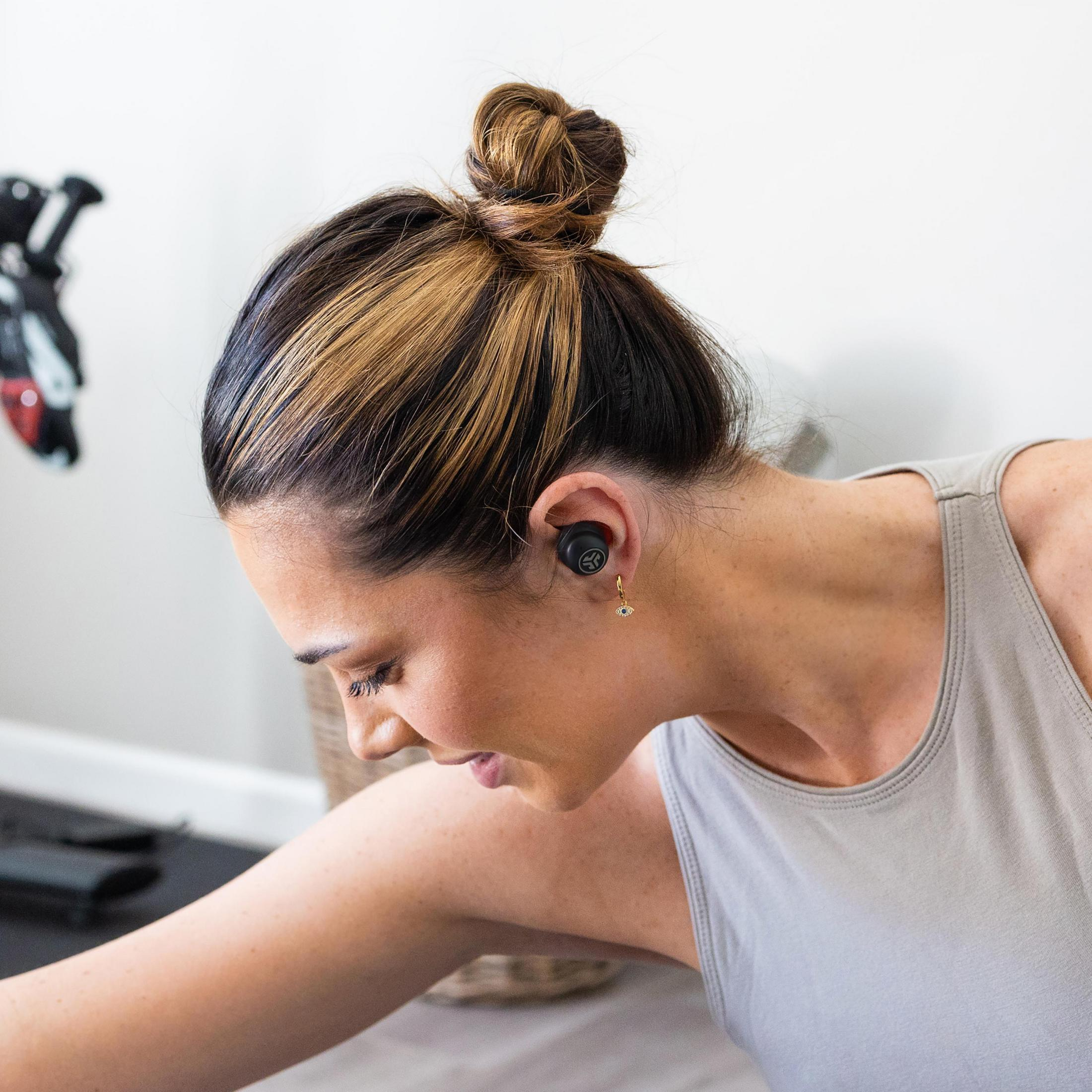 Kobieta ze słuchawką w uchu, patrząca w dół. Włosy są spięte w kok. Tło to pokój.