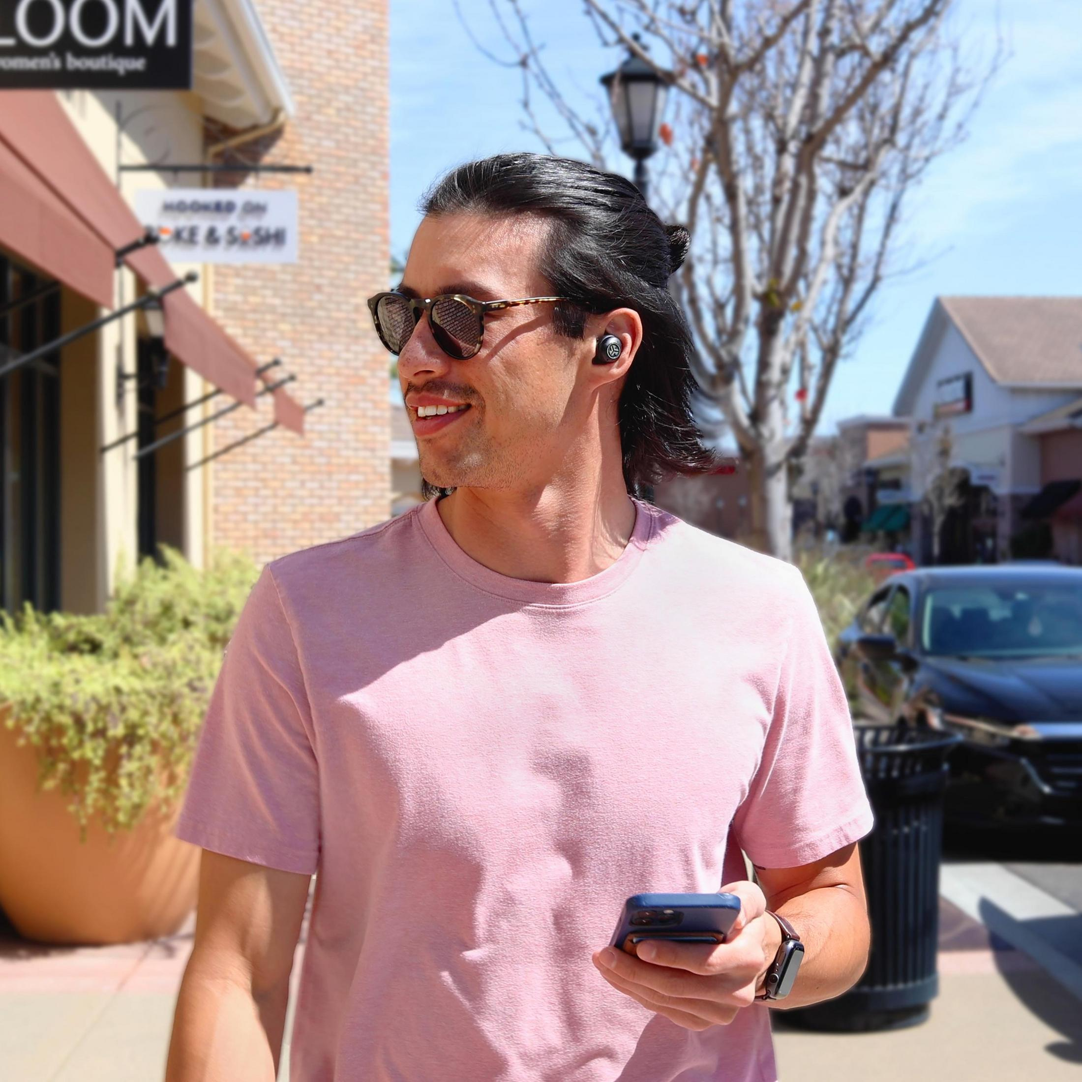 Mężczyzna w różowej koszuli, słuchawkach, okularach przeciwsłonecznych, telefonie, idący chodnikiem.