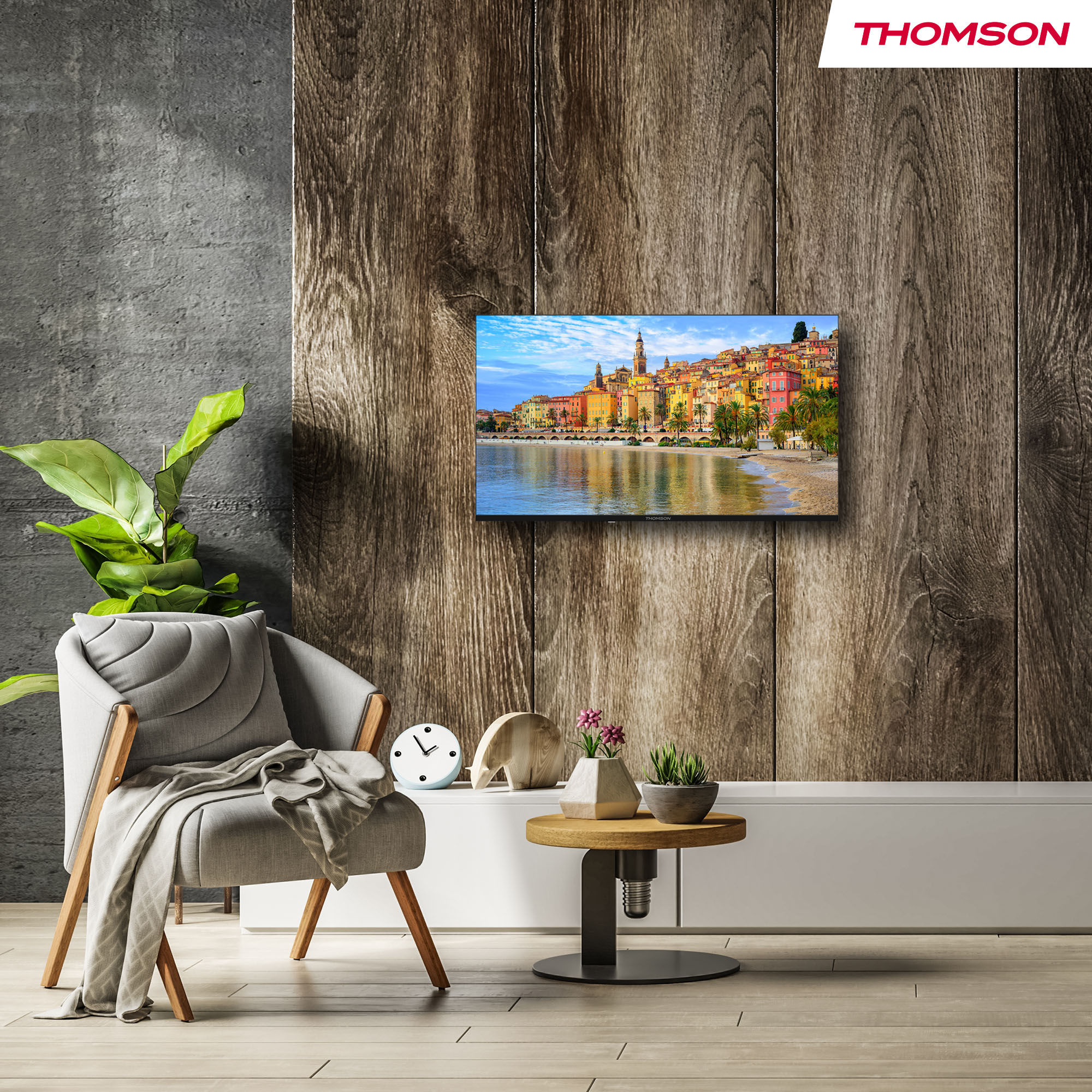 Oprawiony obraz kolorowego nadmorskiego miasteczka. Wisi na drewnianej ścianie obok szarego krzesła.