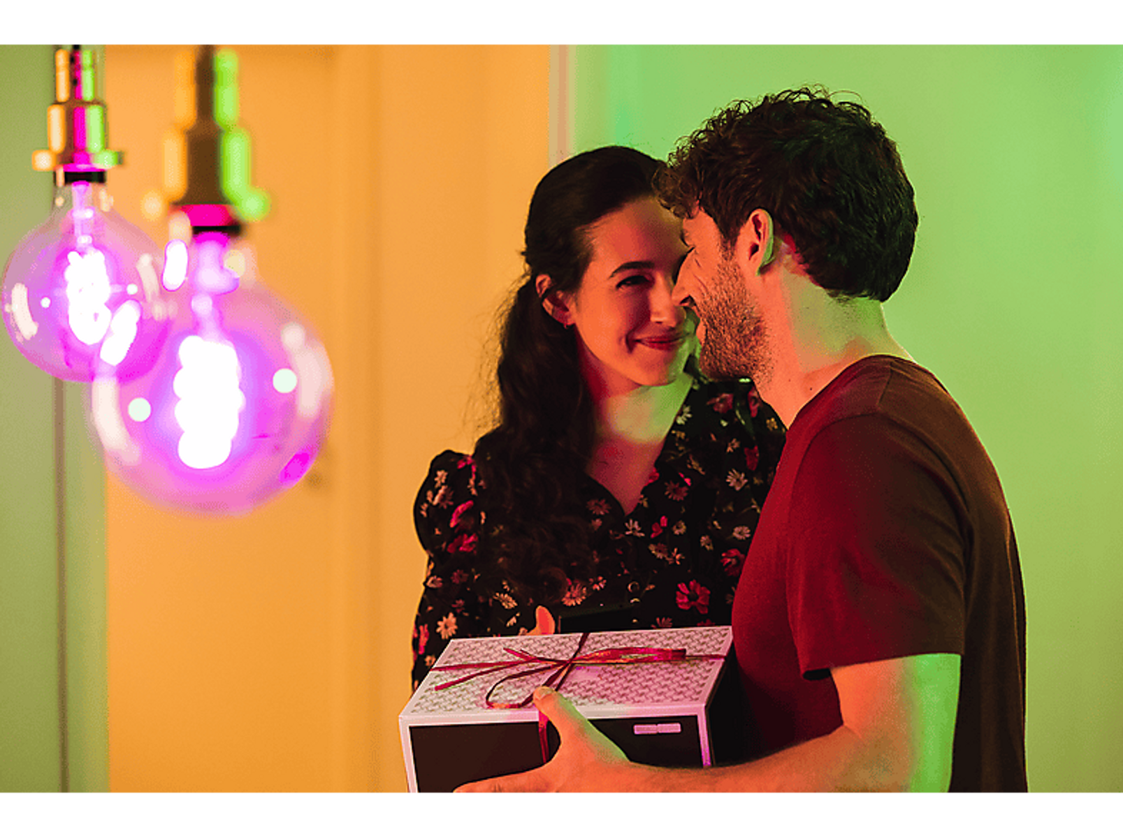 Para w pomieszczeniu, mężczyzna trzymający pudełko z prezentem, patrzy na kobietę, która się uśmiecha.