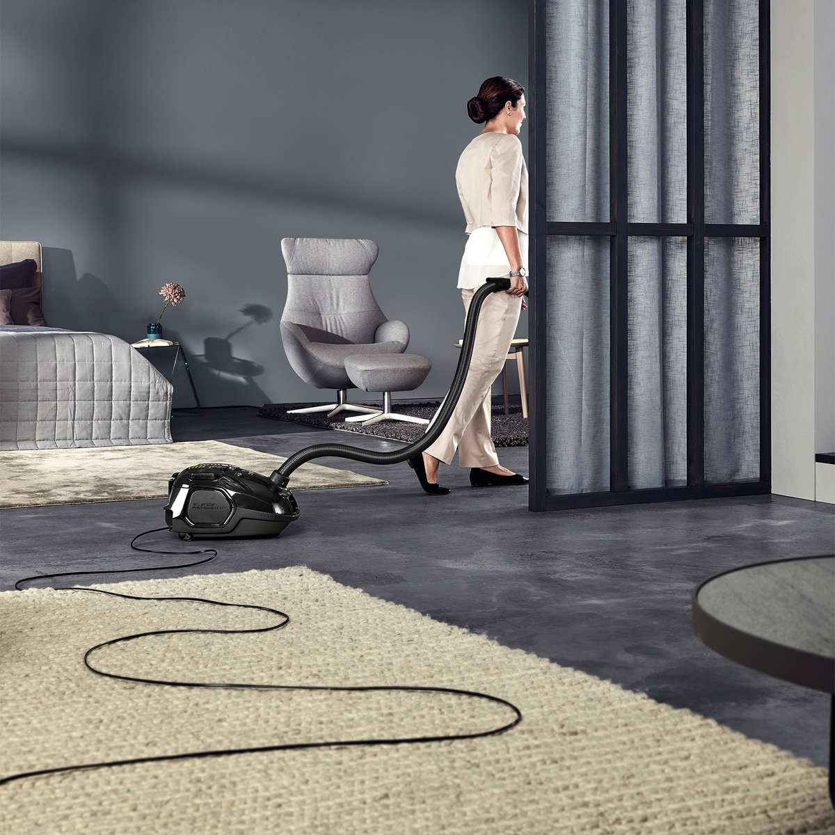Mujer aspira una habitación. Aspiradora en la alfombra. Sillón gris y cama en el fondo.