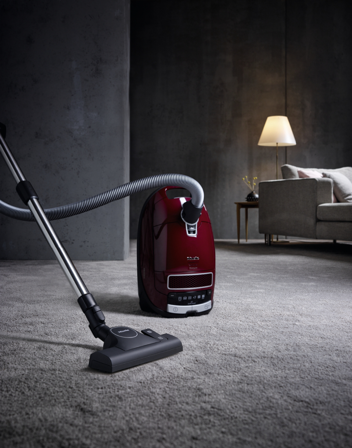 Ein roter Staubsauger und Schlauch auf Teppich, mit Sofa und Lampe im Hintergrund.