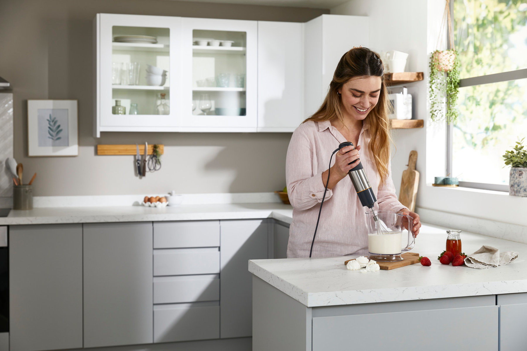 Een vrouw in een keuken gebruikt een staafmixer om ingrediënten in een glazen pot te mengen.