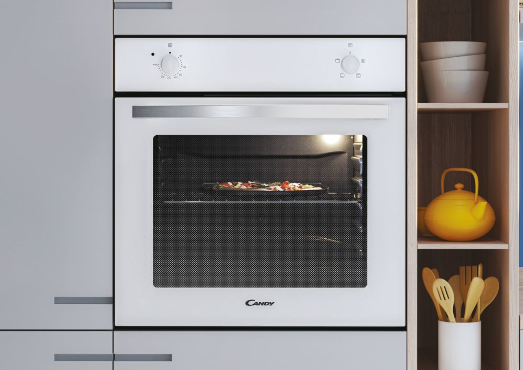 Forno bianco con pizza dentro. Mensola in legno con ciotole e teiera gialla. Armadi grigi.