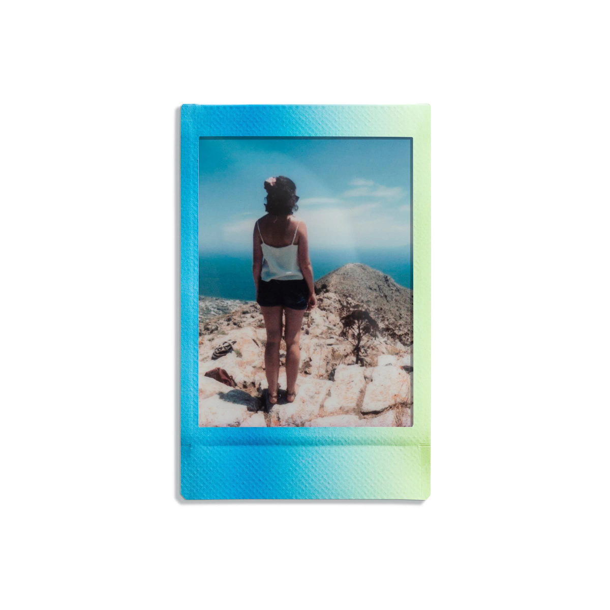 Foto Polaroid: una mujer está en rocas, mirando al mar. Cielo azul.