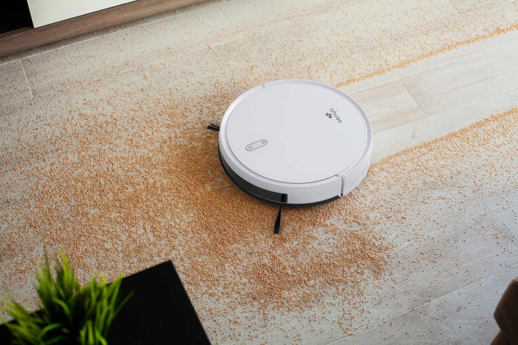 Weißer Roboter-Staubsauger auf einem Holzboden, der mit Körnern übersät ist.