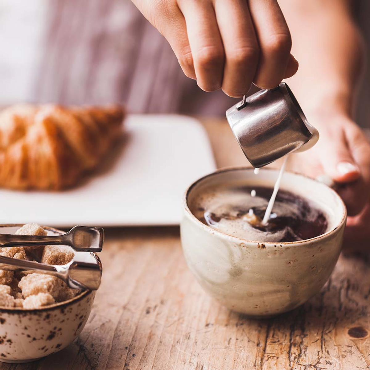 Ręka wlewa śmietankę do filiżanki kawy z ciastem i cukierniczką.