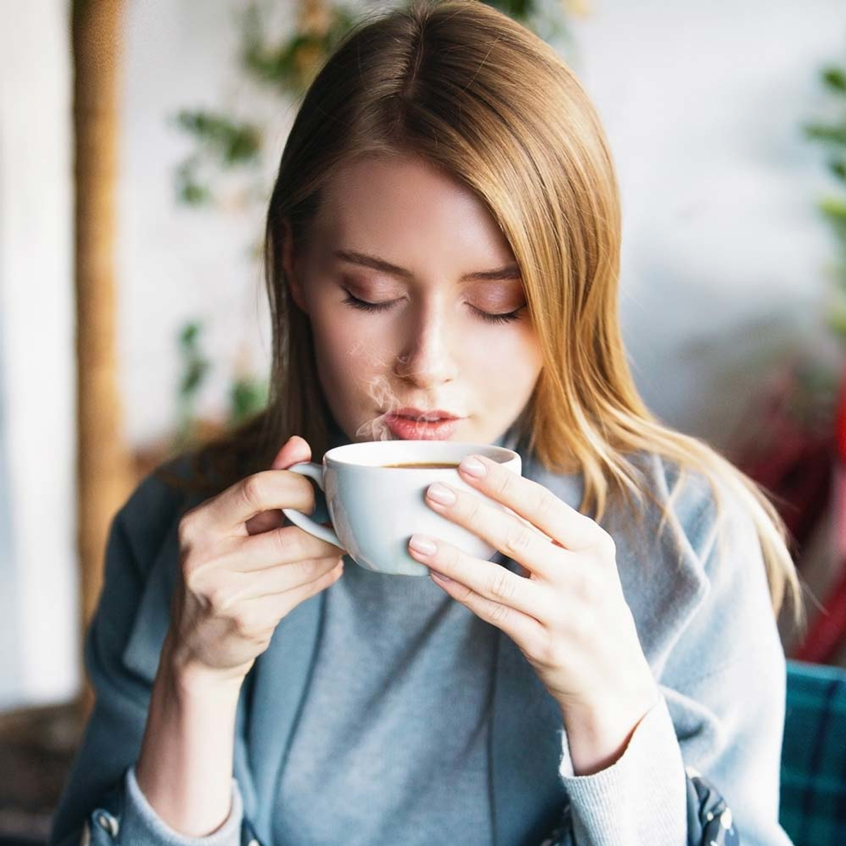 Kobieta trzymająca białą filiżankę kawy, para unosi się, oczy zamknięte. Miękkie oświetlenie.