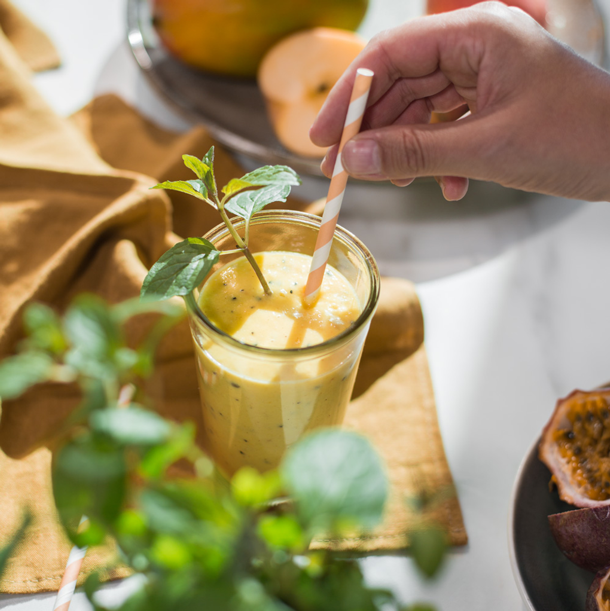 Eine Hand hält einen Strohhalm in einem Smoothie-Glas. Obst, Minze und eine gelbe Serviette sind dabei.