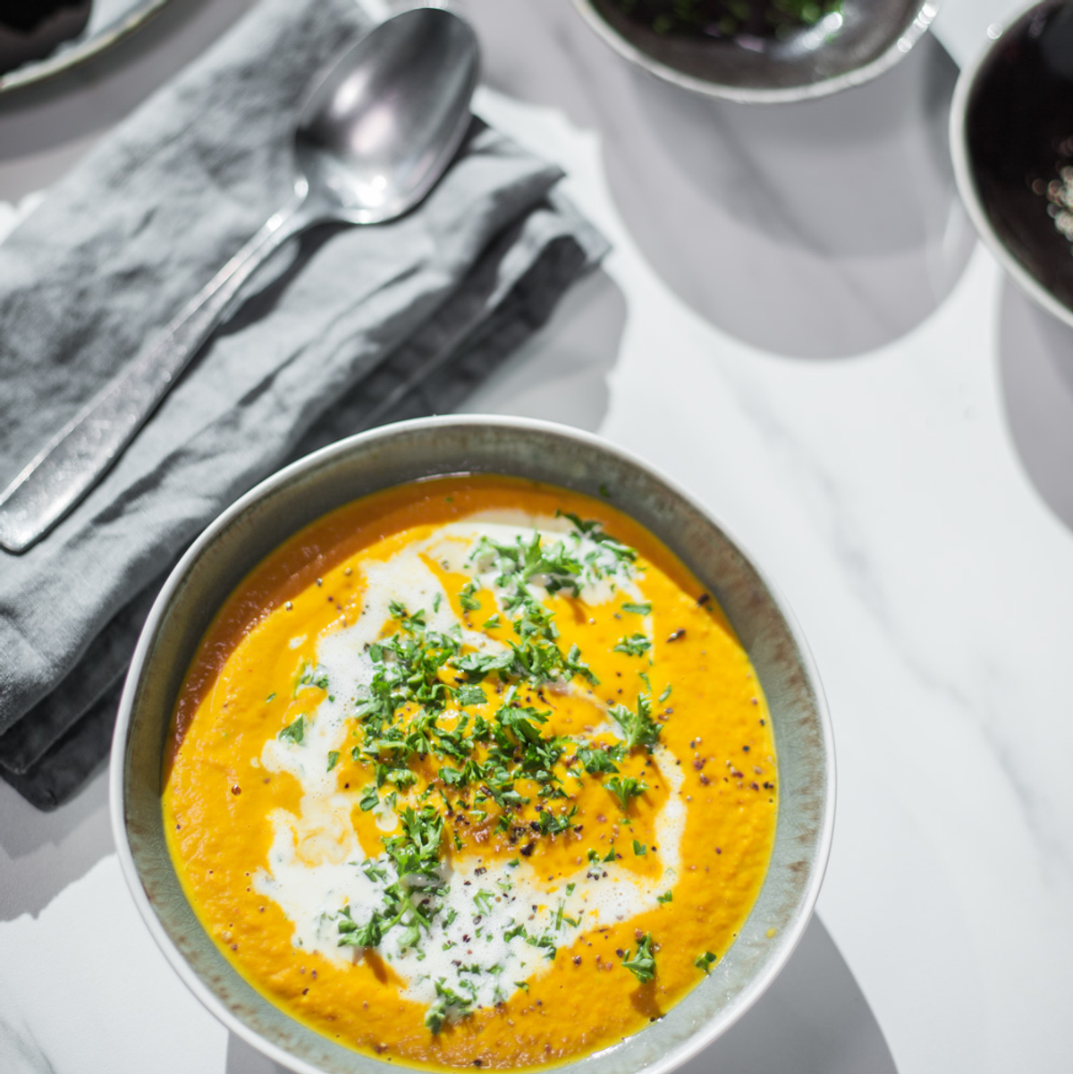 Orangene Suppe in Keramikschale mit Sahnewirbel und Petersilie, Marmoroberfläche.