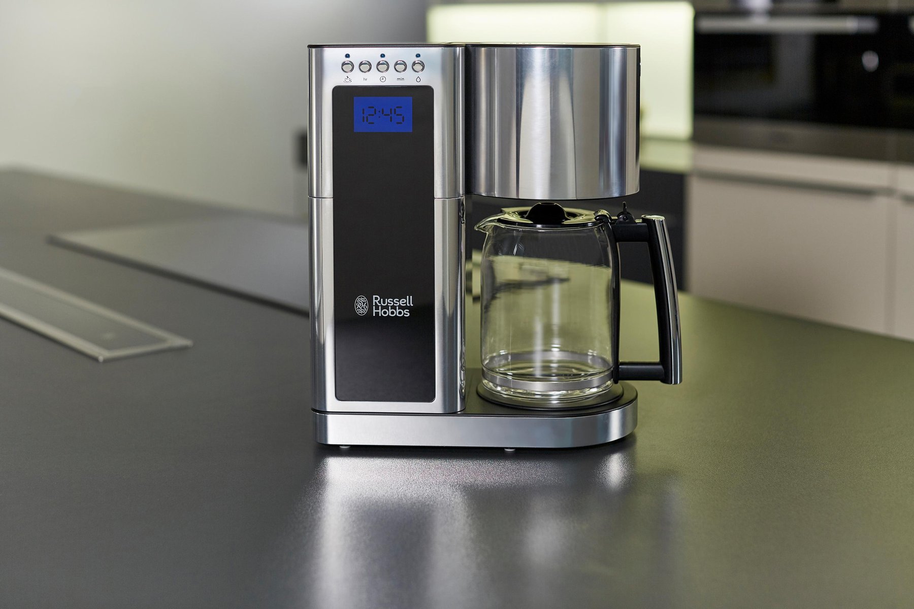 Macchina per caffè in acciaio inox su un piano grigio. Visualizza l'ora 12:45. Brocca di vetro a destra.