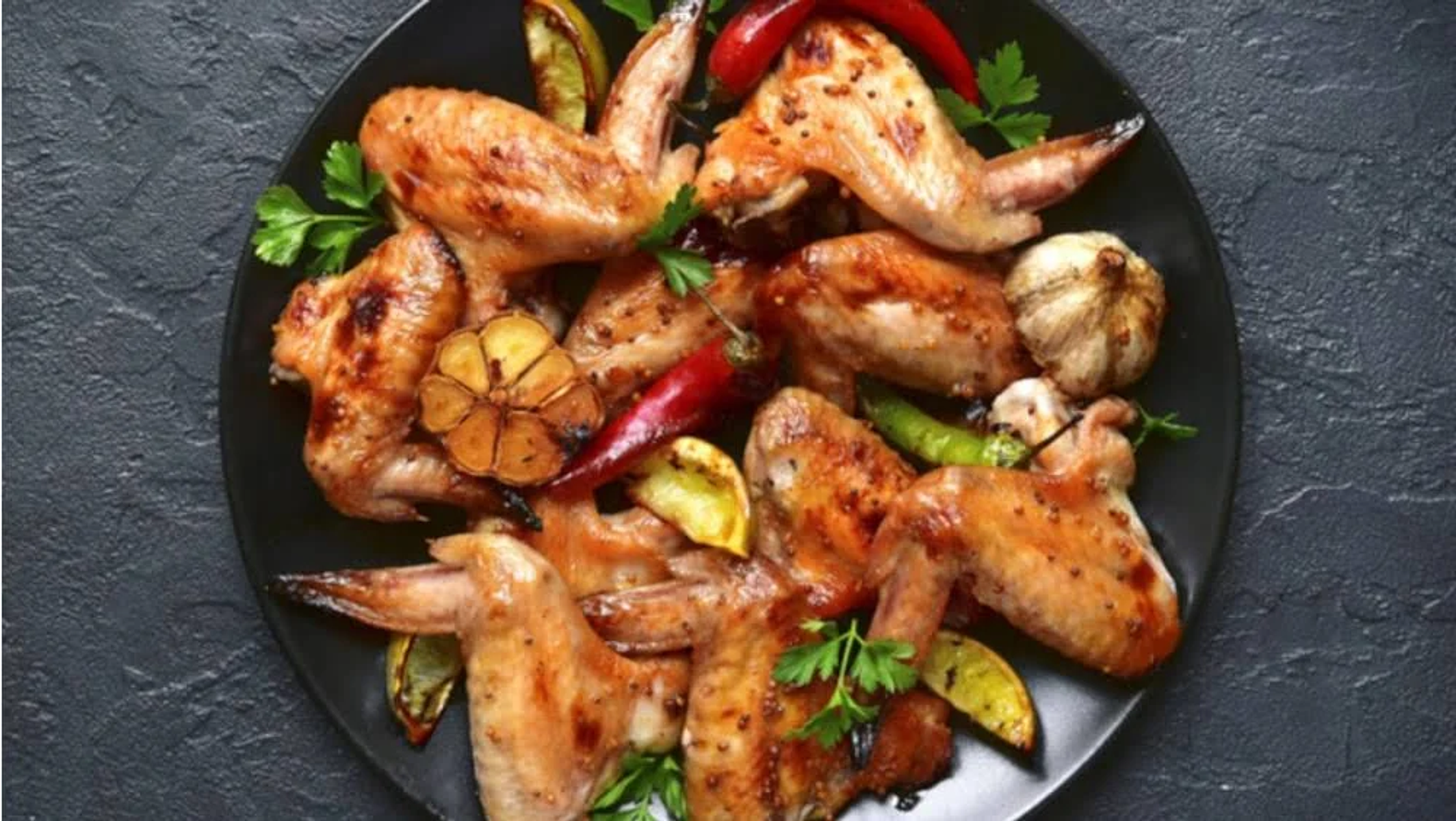 Draufsicht auf gebratene Hähnchenflügel auf einem schwarzen Teller, garniert mit Kräutern, Zitrone und Chili.