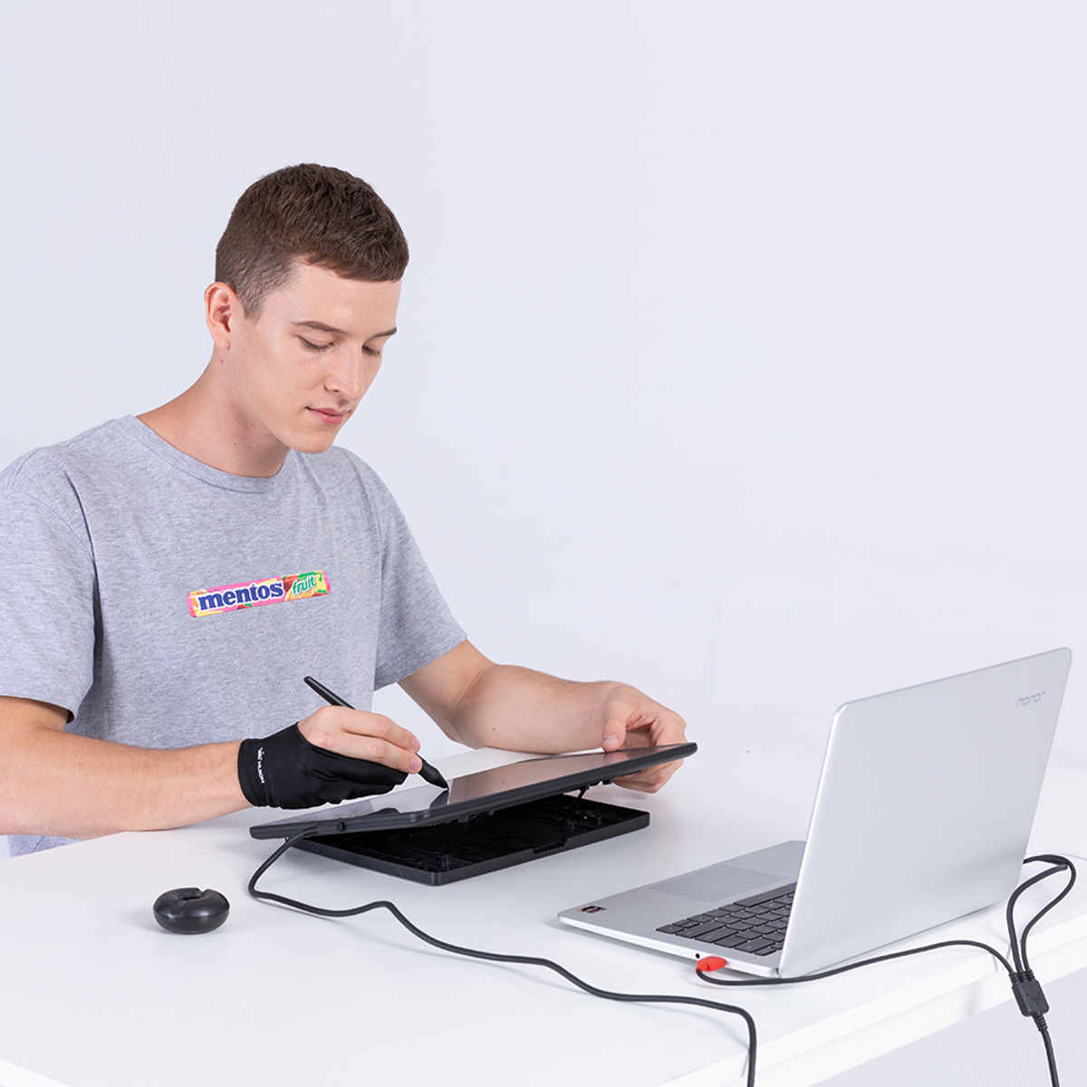 A man using a laptop and a graphics tablet, wearing a black glove, and a grey shirt.