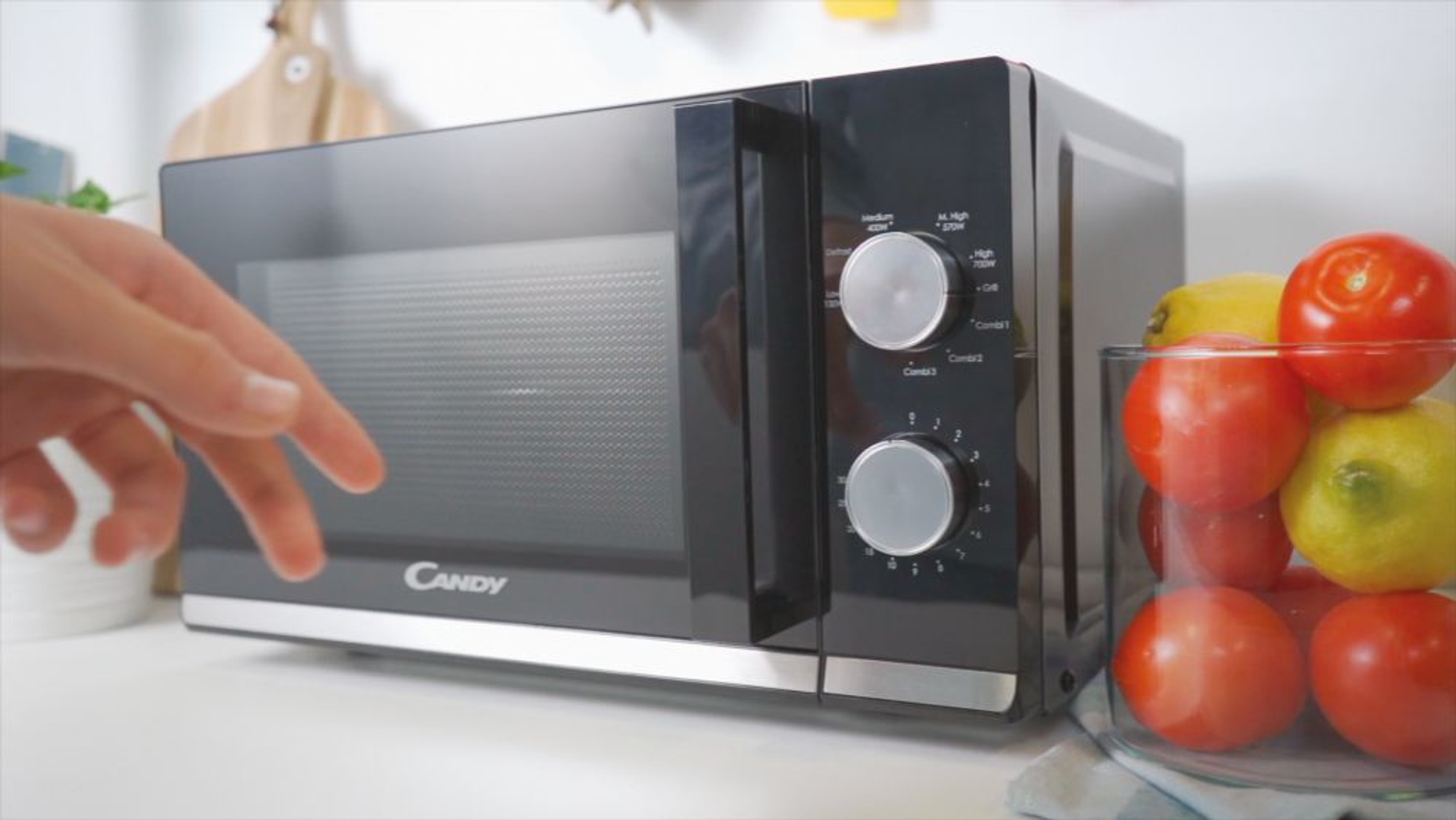 Czarna kuchenka mikrofalowa ze szklanymi drzwiami. Ręka dotyka kuchenki mikrofalowej. Widoczne pomidory i cytryny.