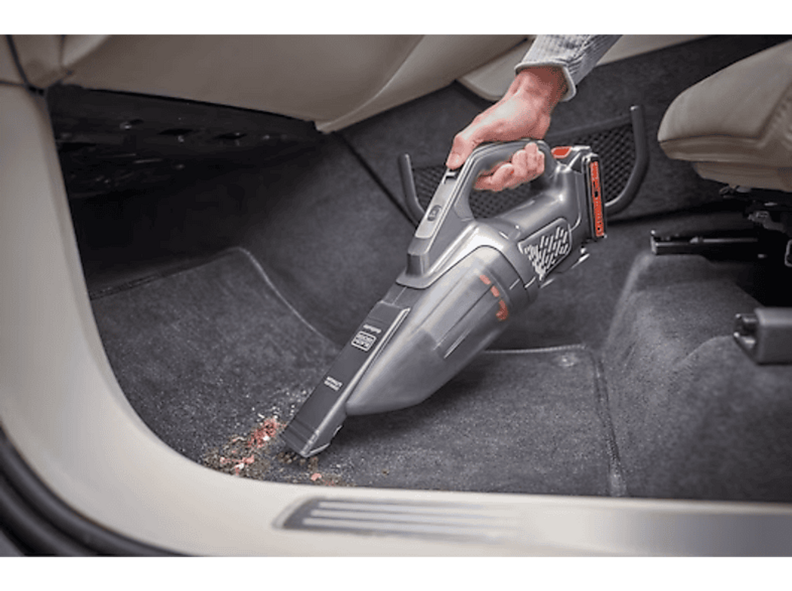 Una persona aspirando el interior de un coche con una aspiradora de mano. Se ve suciedad y migas en la alfombra.