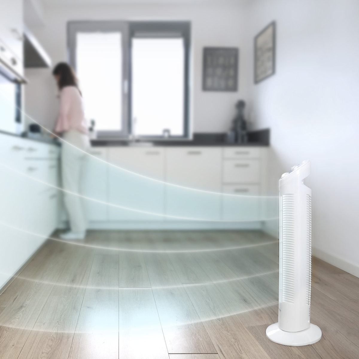 White fan on a wooden floor, with a person in the blurry background in a kitchen setting.