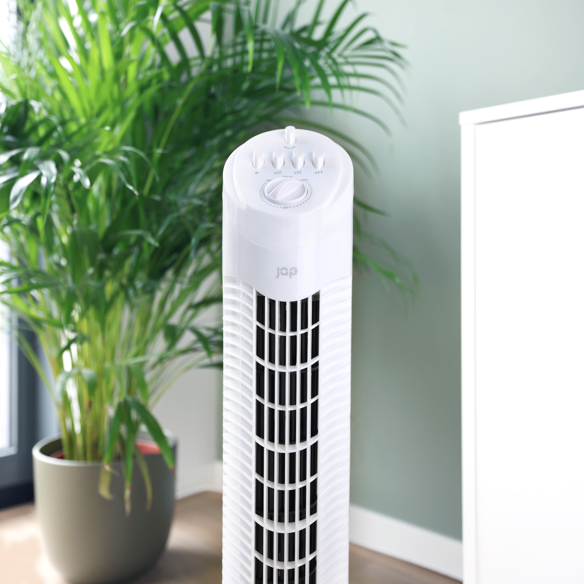 White tower fan with controls, a potted plant, and a white cabinet. Green wall backdrop.