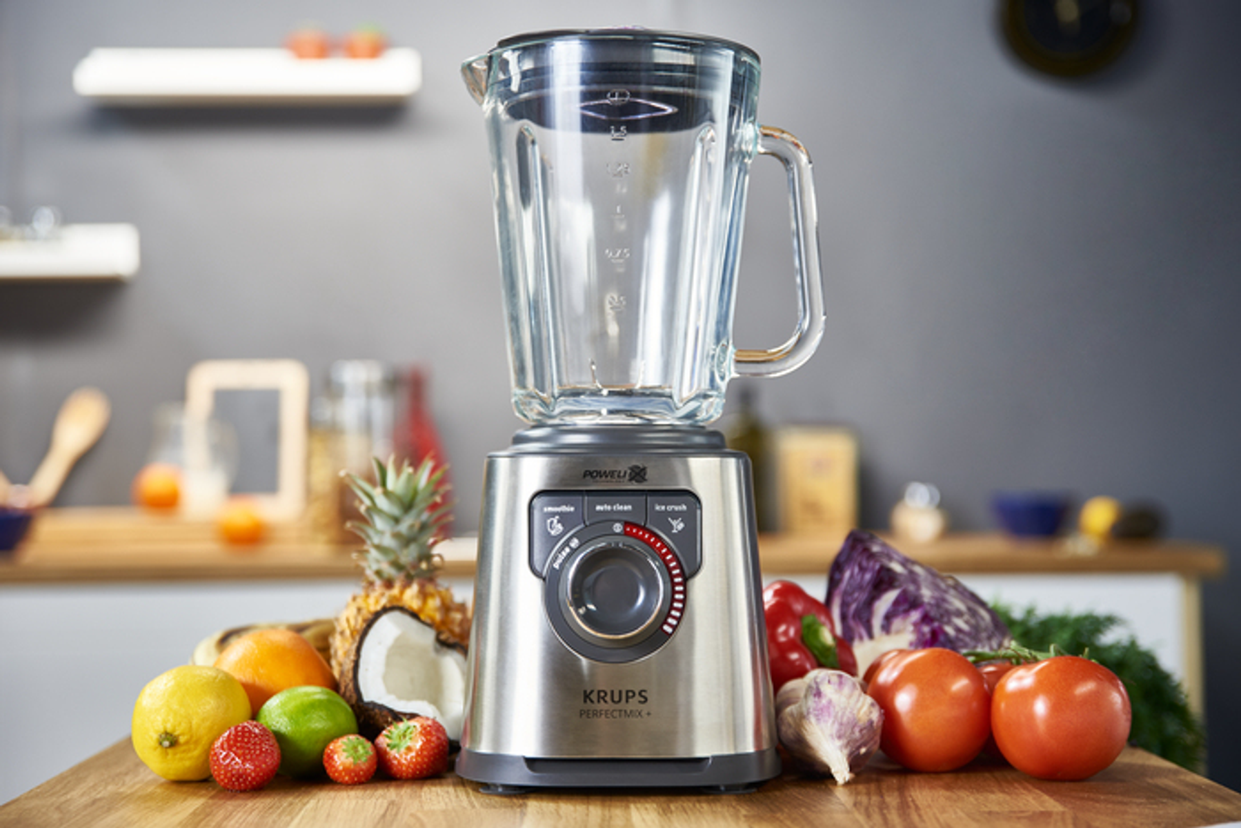 Ein Krups-Blender mit Früchten auf einem Holztisch vor grauem Hintergrund. Der Mixer ist im Fokus.