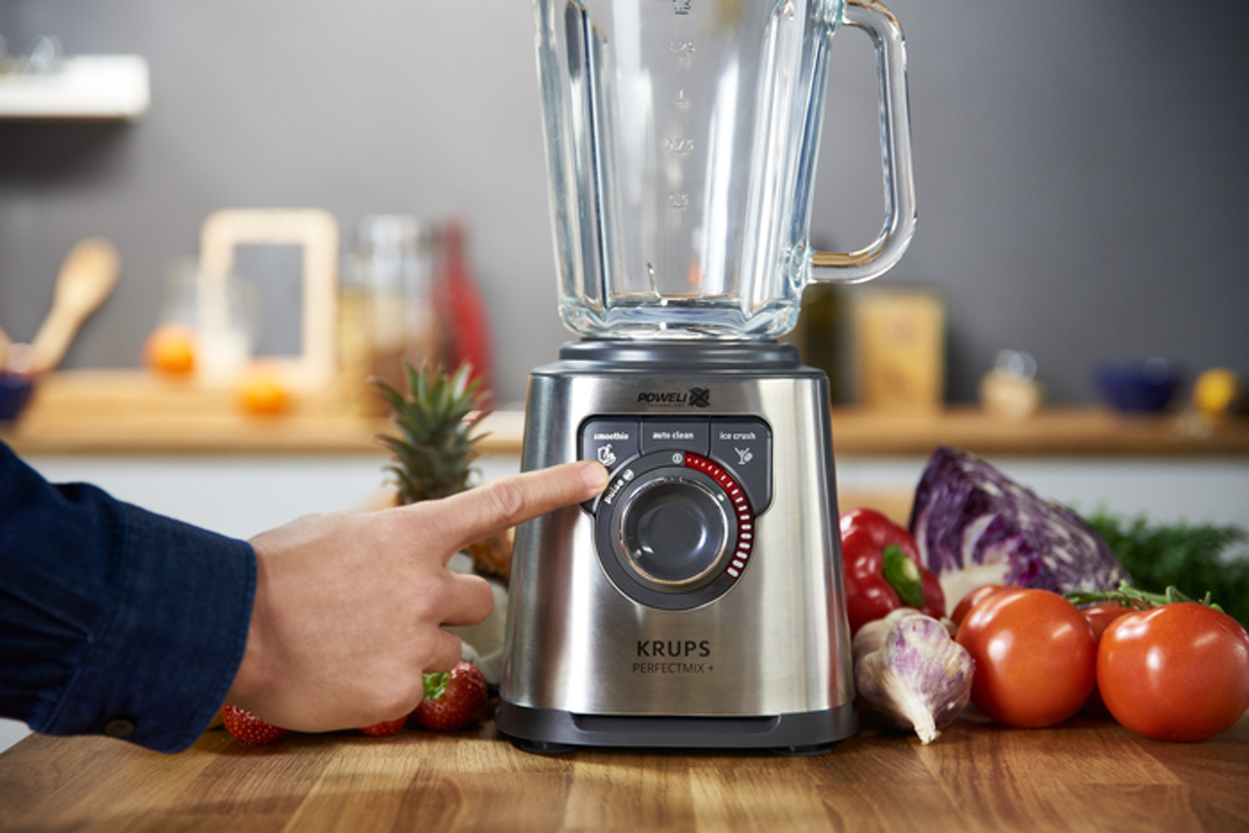 Die Hand einer Person dreht einen Drehregler an einem silbernen Mixer. Gemüse und Obst umgeben den Mixer.