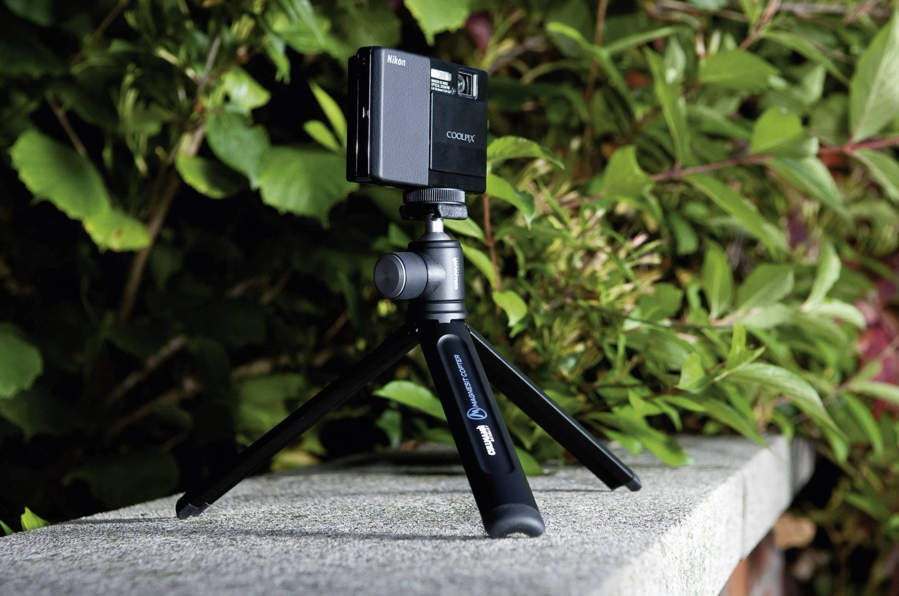 Een zwarte camera op een klein statief, geplaatst op een muur met groene planten op de achtergrond.