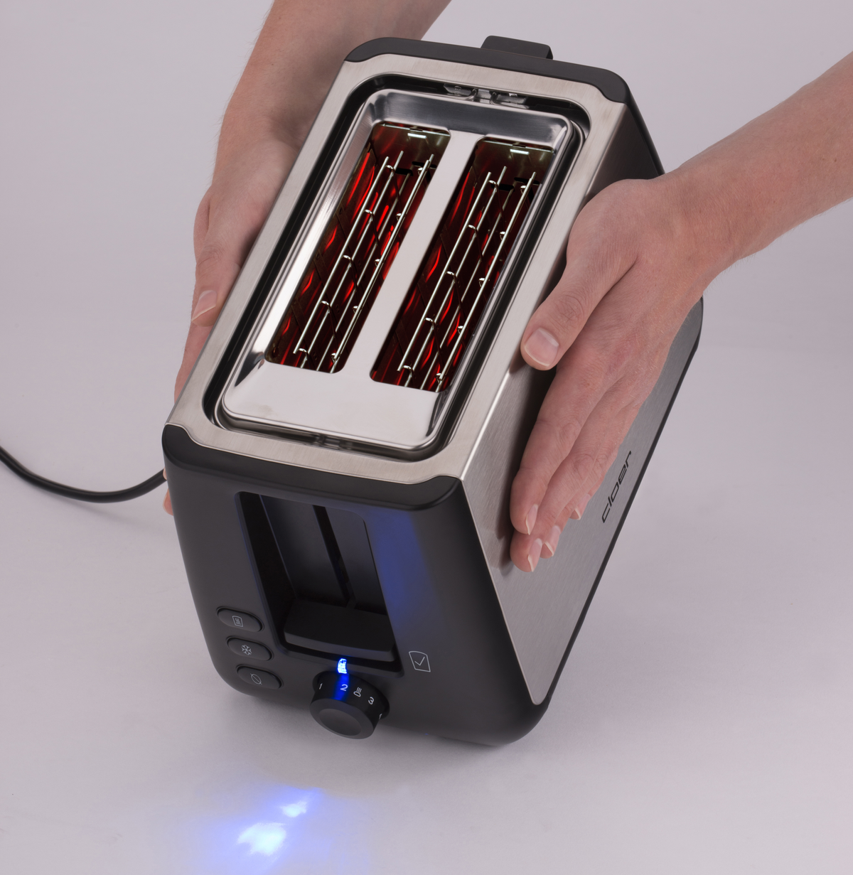 Toster ze srebrnymi bokami i czarnym przodem. Dłonie trzymają go. Białe tło.