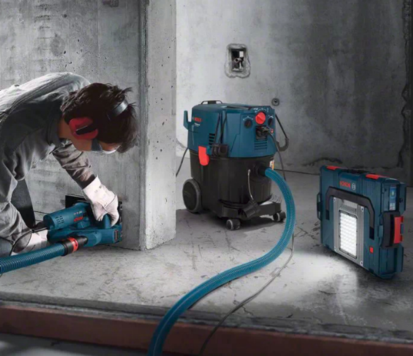 Hombre usando herramienta azul Bosch, aspiradora y luz en habitación de concreto.
