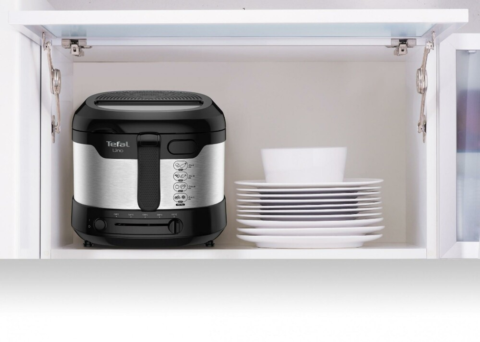 Frytkownica Tefal Uno w białej szafce, obok stosu białych talerzy.