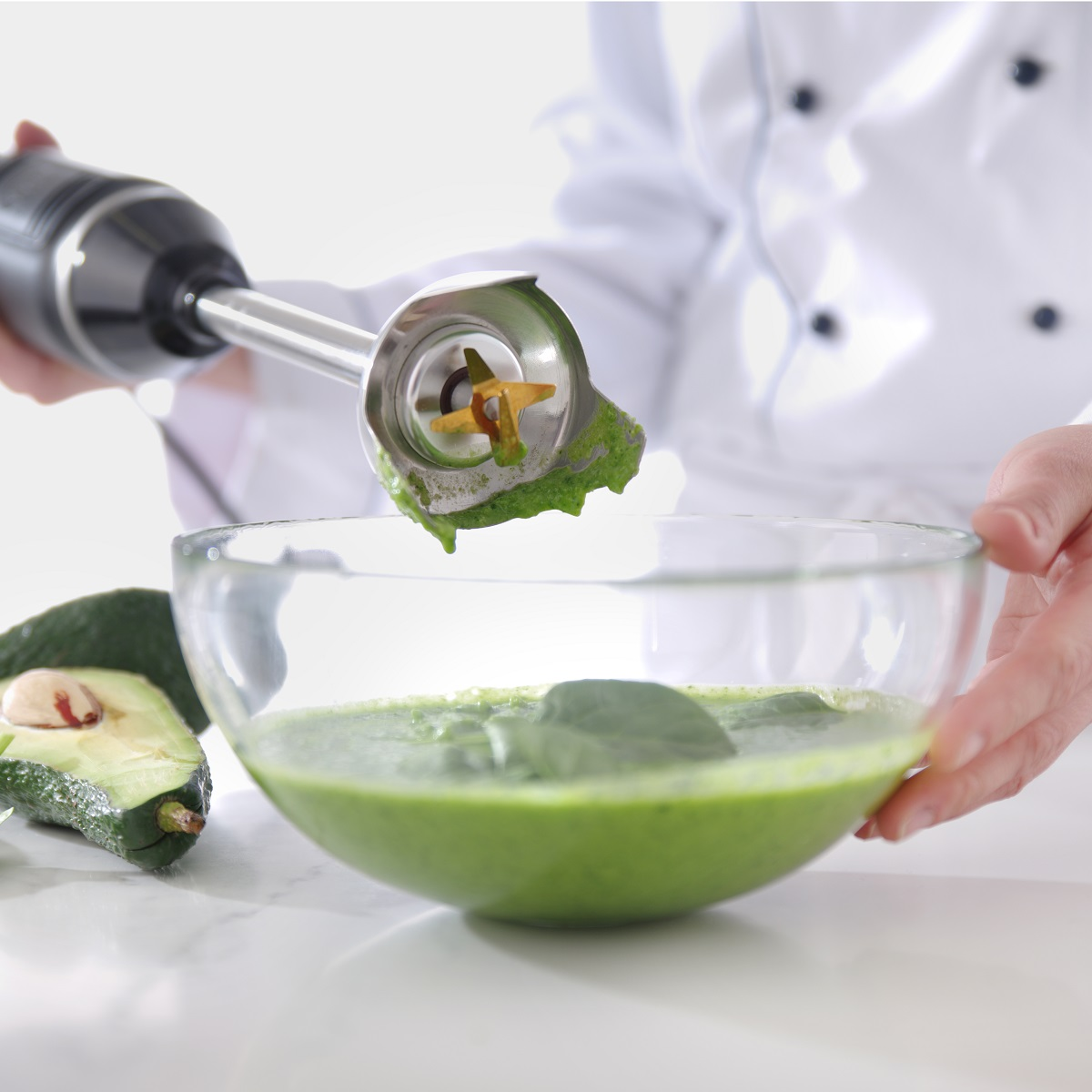 Eine Person verwendet einen Stabmixer, um grüne Suppe in einer Glasschüssel mit Avocadoscheiben zu mixen.