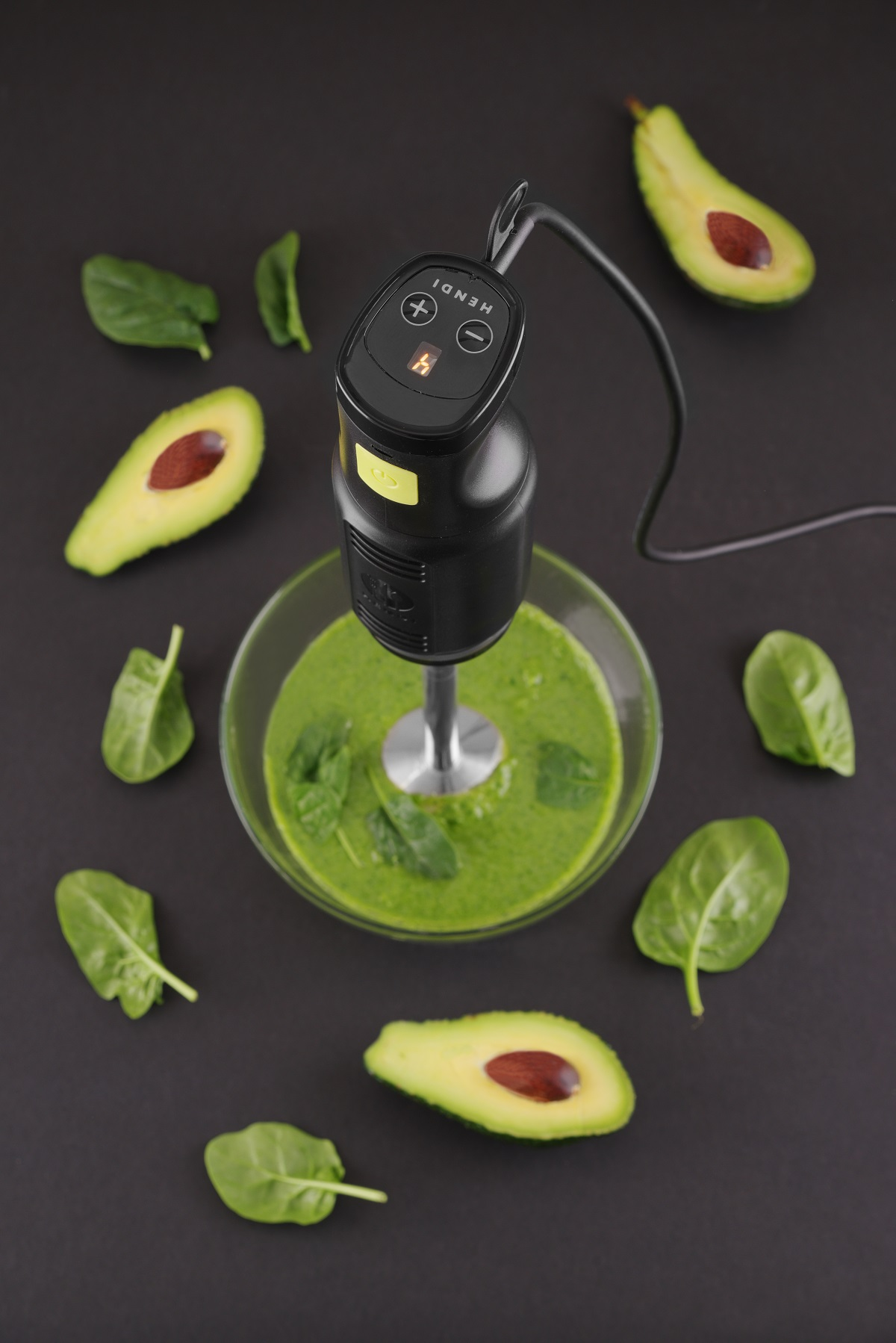 Stabmixer mit grüner Mischung in einer Glasschüssel. Avocado und Spinat umgeben die Schüssel.