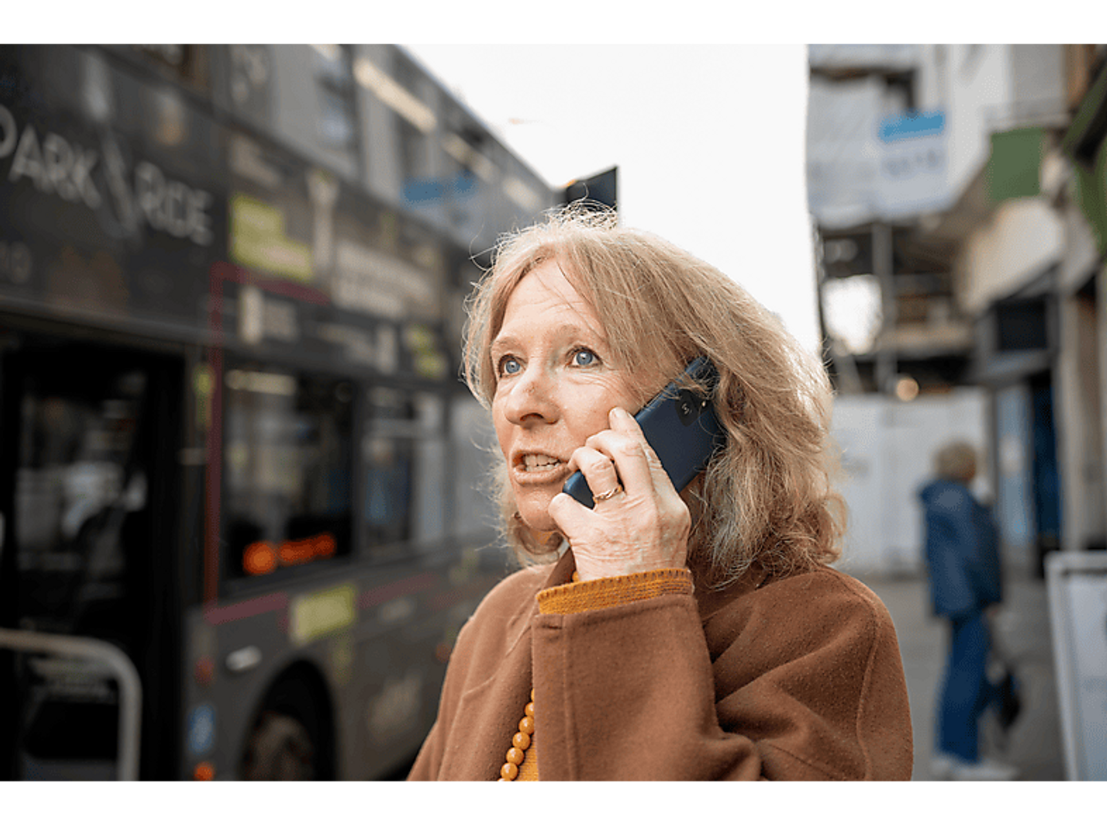Frau telefoniert in der Nähe eines Busses und Gebäuden. Sie trägt einen braunen Mantel.