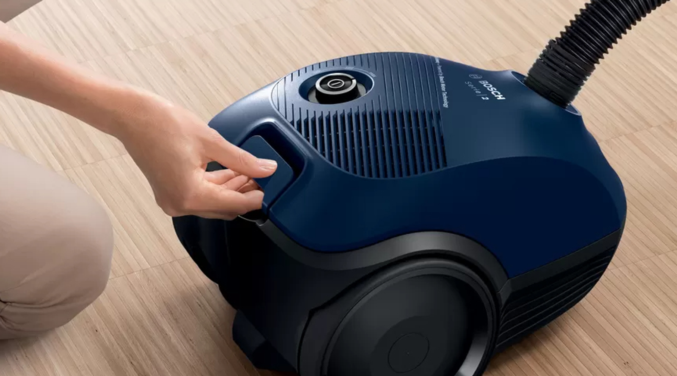 Ręka otwiera boczną komorę niebieskiego odkurzacza na drewnianej podłodze.