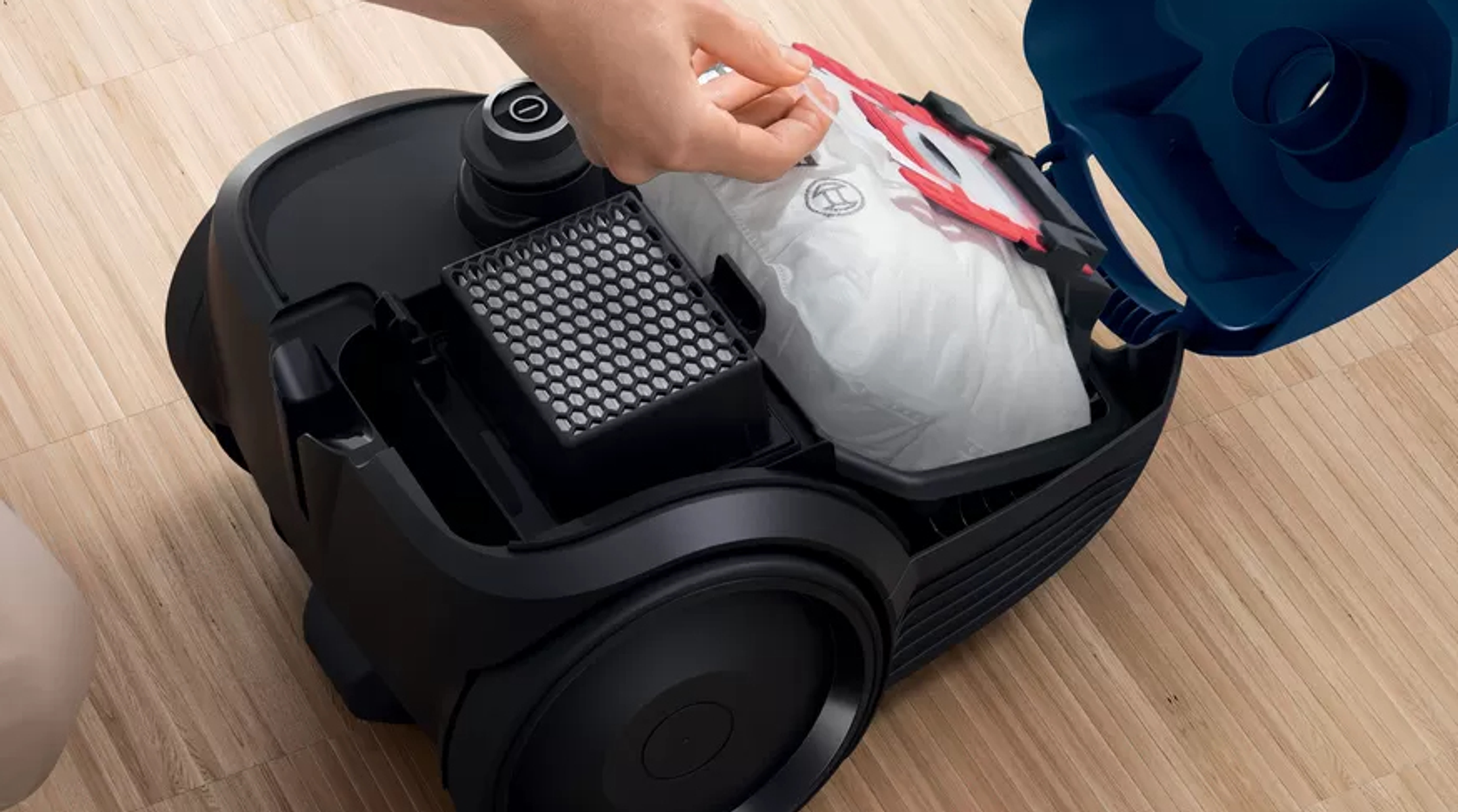 Osoba wkłada biały filtr workowy do czarnego odkurzacza. Drewniane tło.