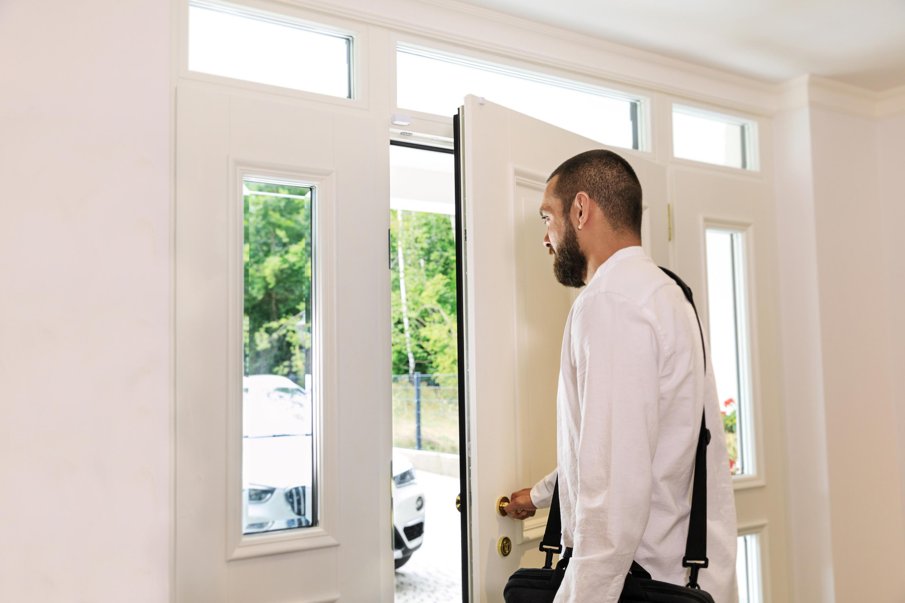 Mężczyzna w białej koszuli otwiera białe drzwi, patrząc na zewnątrz.