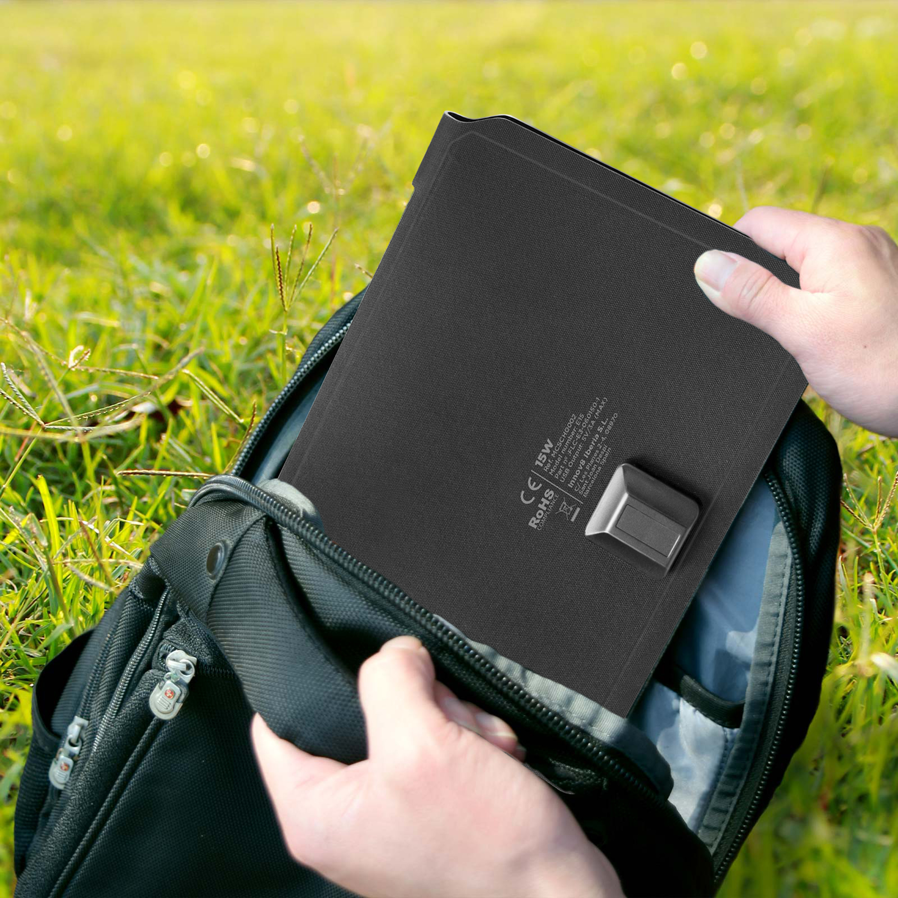 Las manos colocan un panel solar negro en una mochila negra sobre hierba verde.