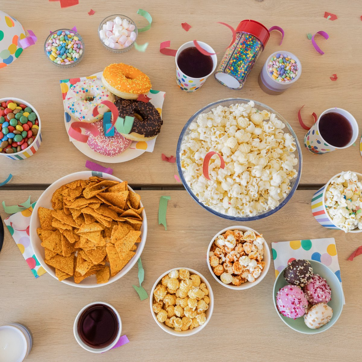 A table with various snacks like donuts, popcorn, chips, and candies, with colorful confetti.