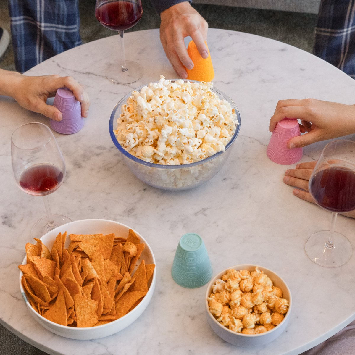 Table with snacks. Popcorn in a large bowl, tortilla chips, and small cups. People's hands reaching for food.