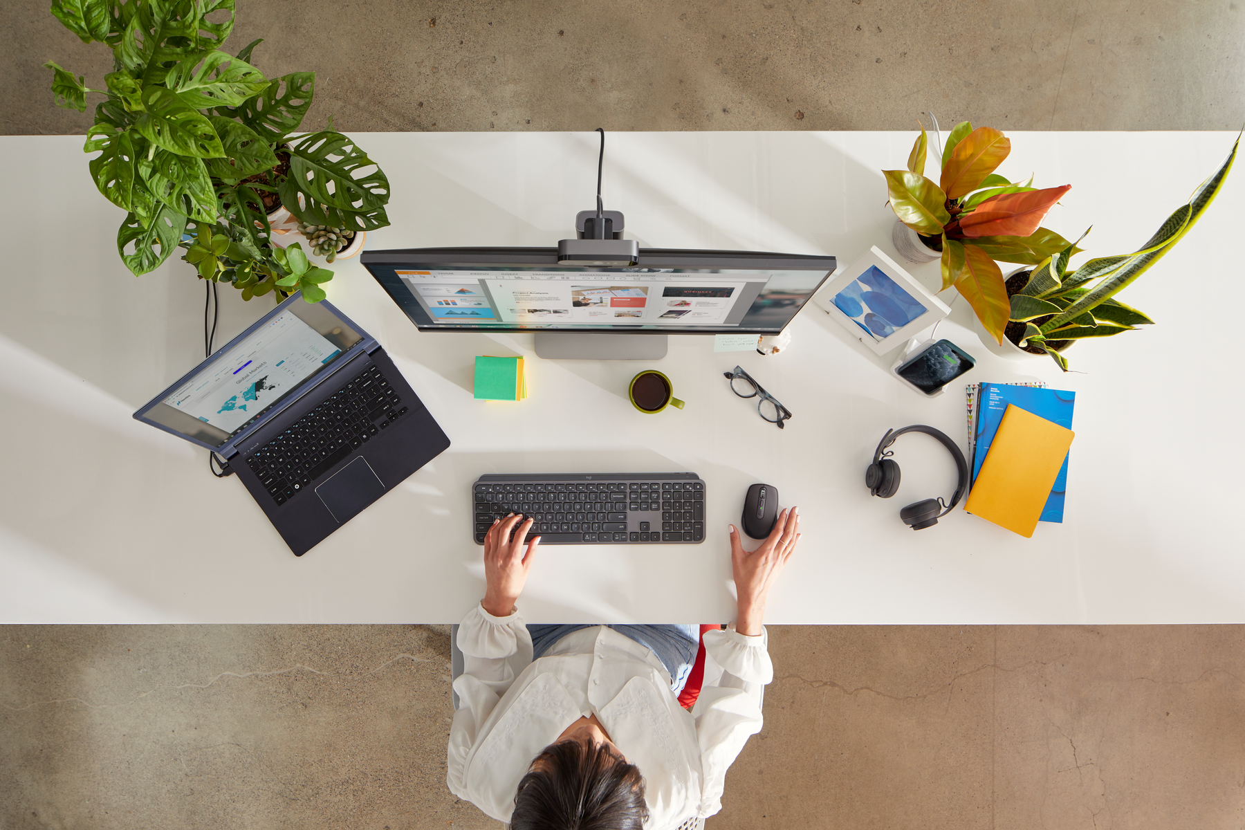 Widok z góry na biurko z monitorem, laptopem, klawiaturą, myszą, roślinami i rękami osoby.