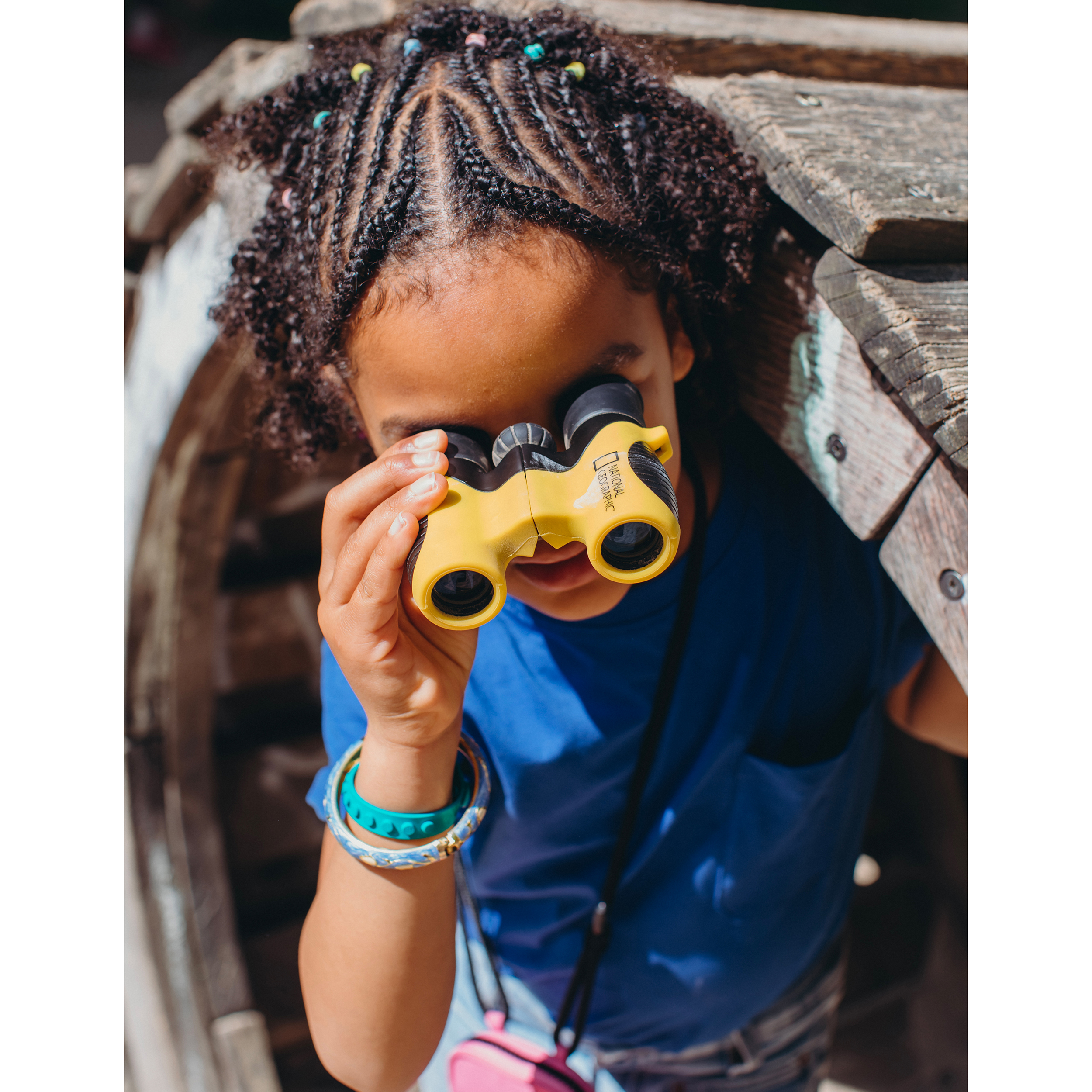 Młoda dziewczyna z warkoczami patrząca przez żółtą lornetkę w drewnianej konstrukcji.