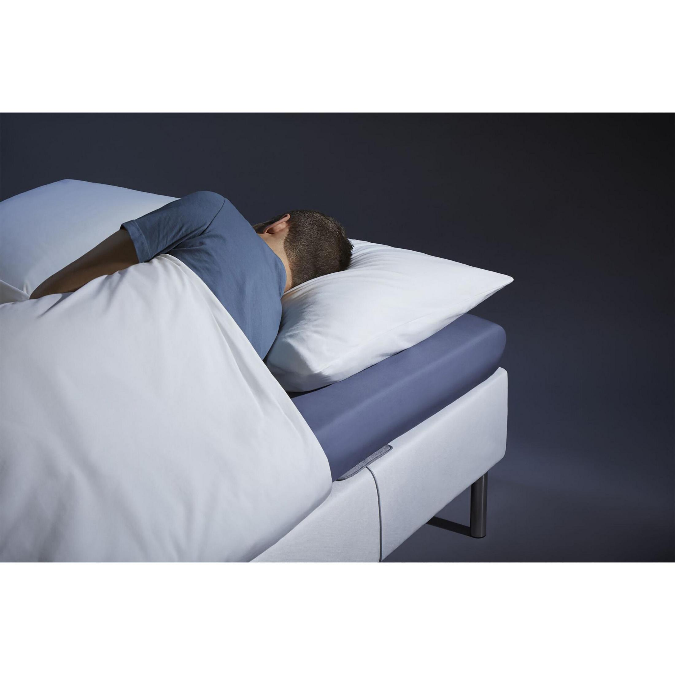 Una persona dorme su un letto coperto da lenzuola bianche. Testa su un cuscino, in un ambiente buio.