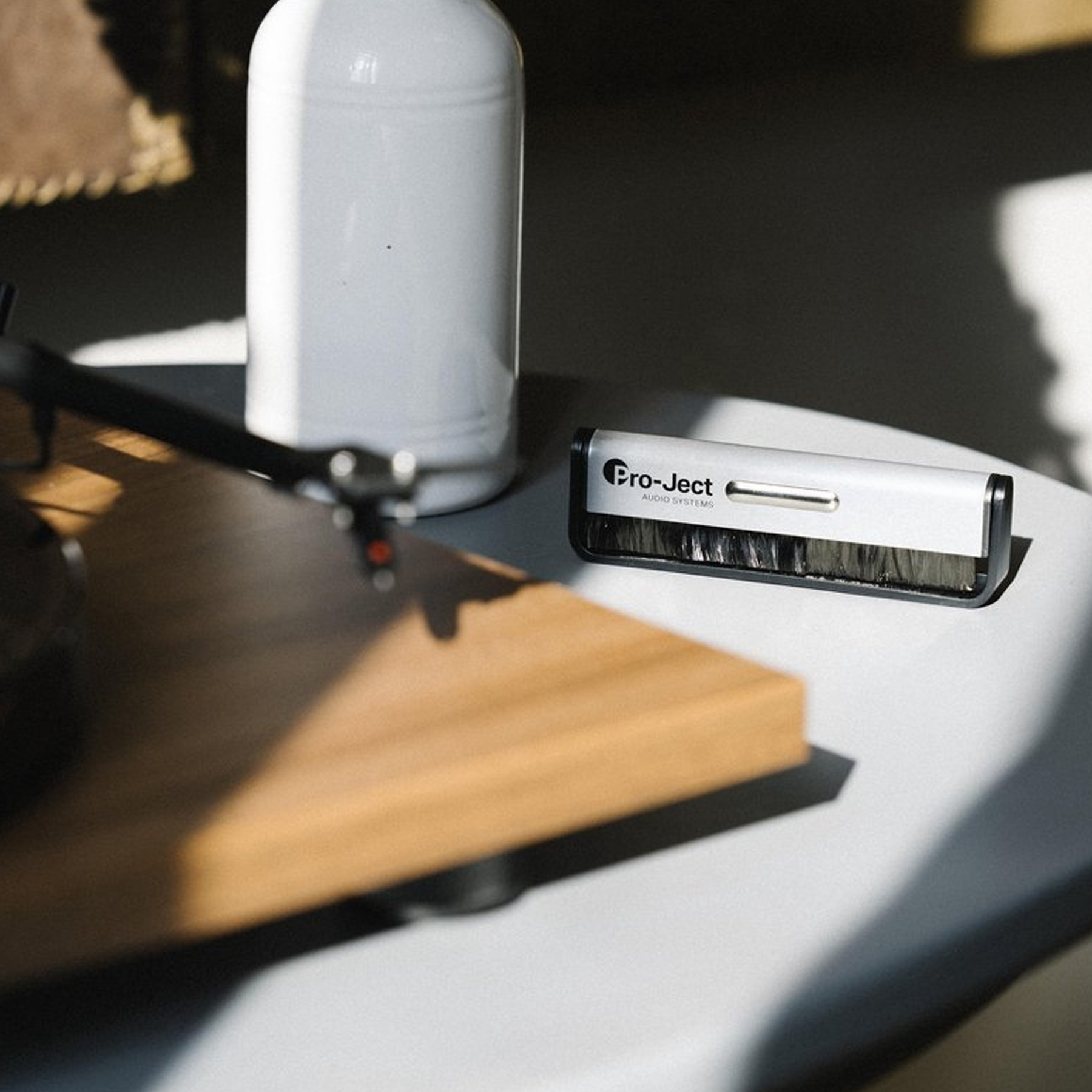 Gramofon ze szczotką Pro-Ject na jasnej powierzchni. Za szczotką biała butelka.
