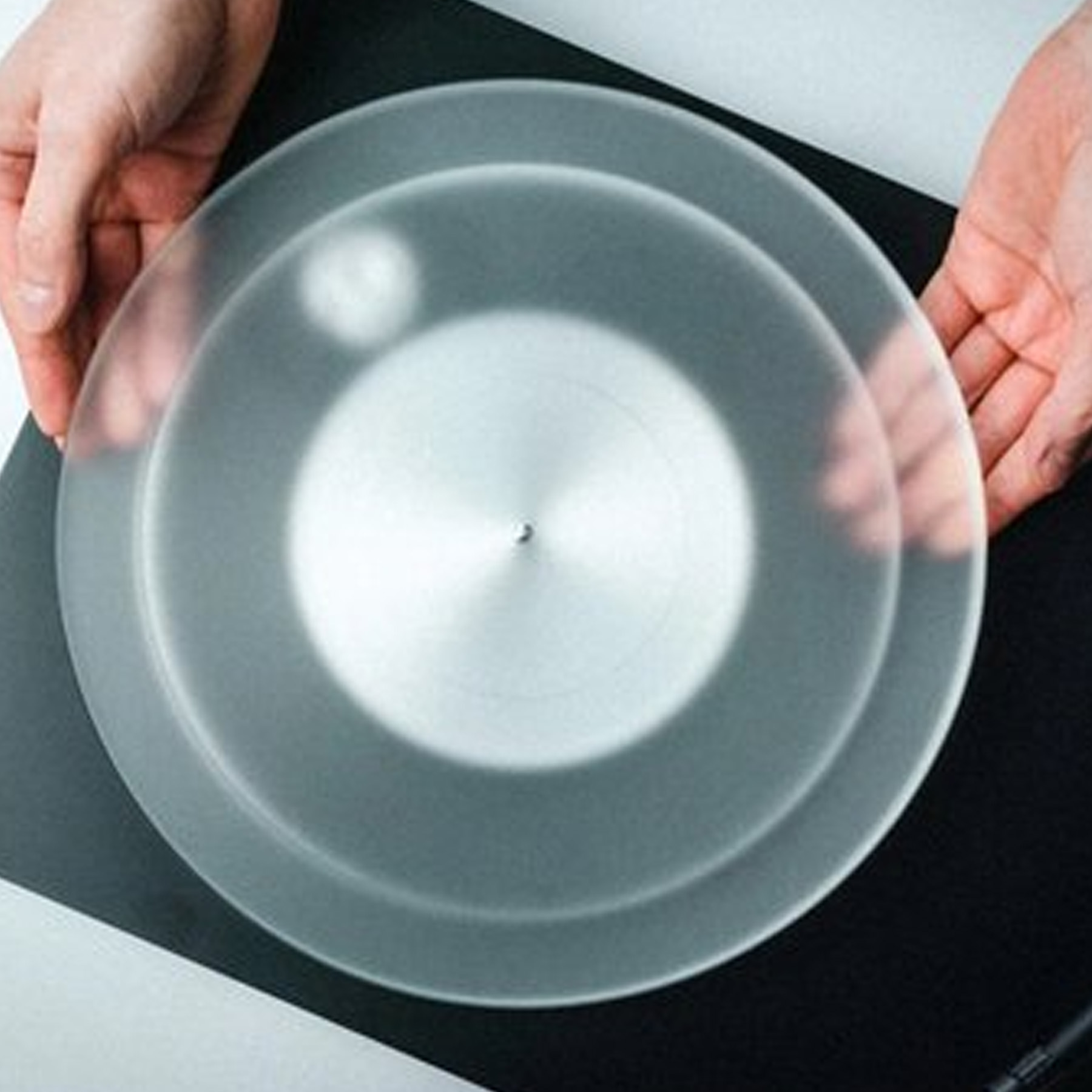 Dwie ręce trzymające przezroczystą, okrągłą osłonę płyty winylowej na czarnej powierzchni.